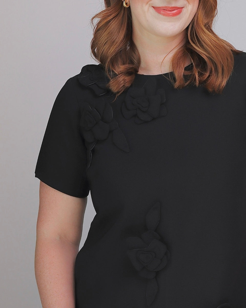 A woman wearing a black short-sleeve dress with 3D floral appliques on the chest and lower skirt, shown against a plain gray background.
