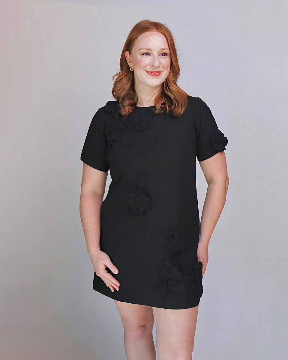 A woman with red hair wearing a black short-sleeve dress with 3D floral appliques, standing against a light gray studio background.