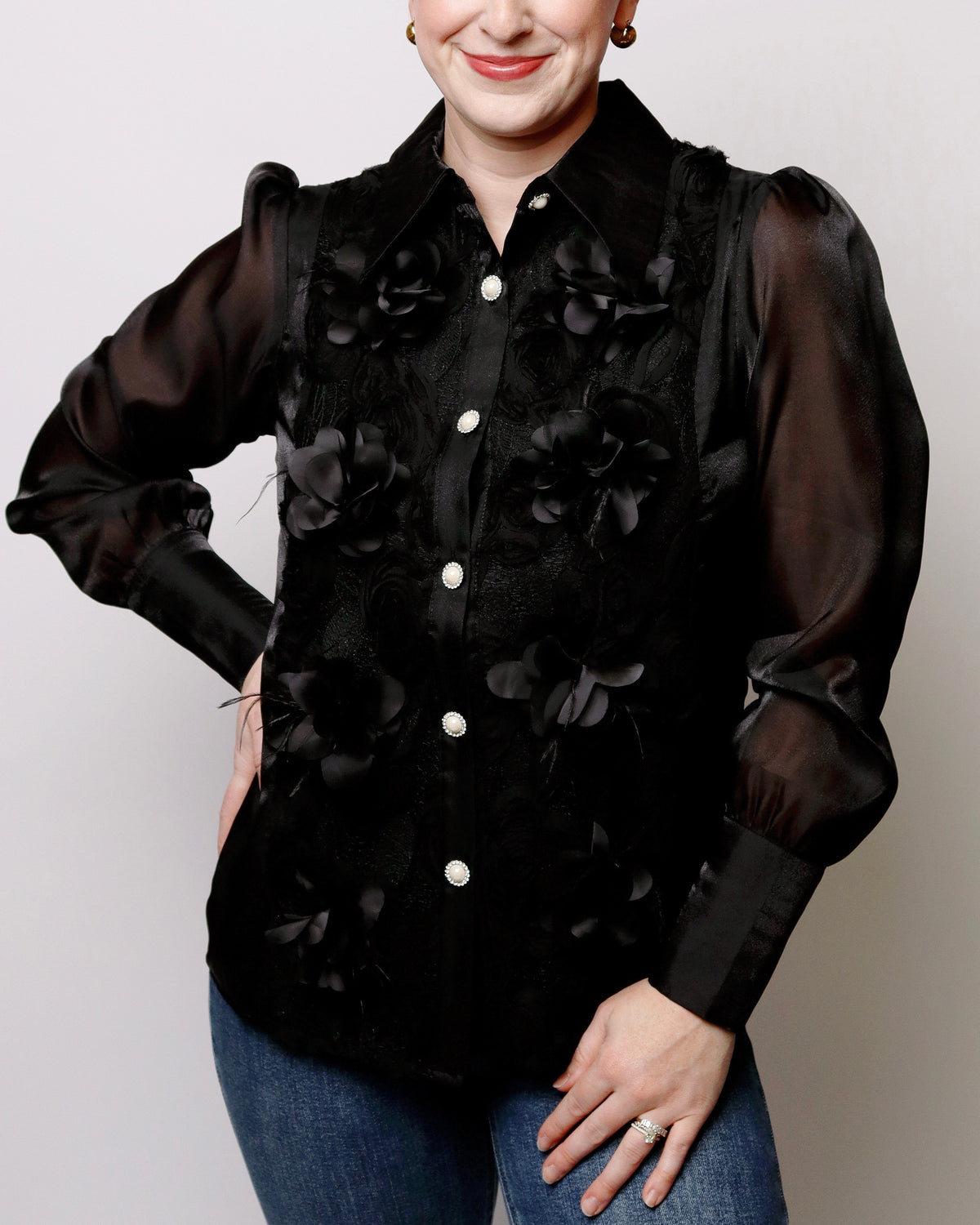 A woman wearing a black long-sleeve blouse with sheer puff sleeves and 3D floral applique, paired with blue jeans, against a light gray background.