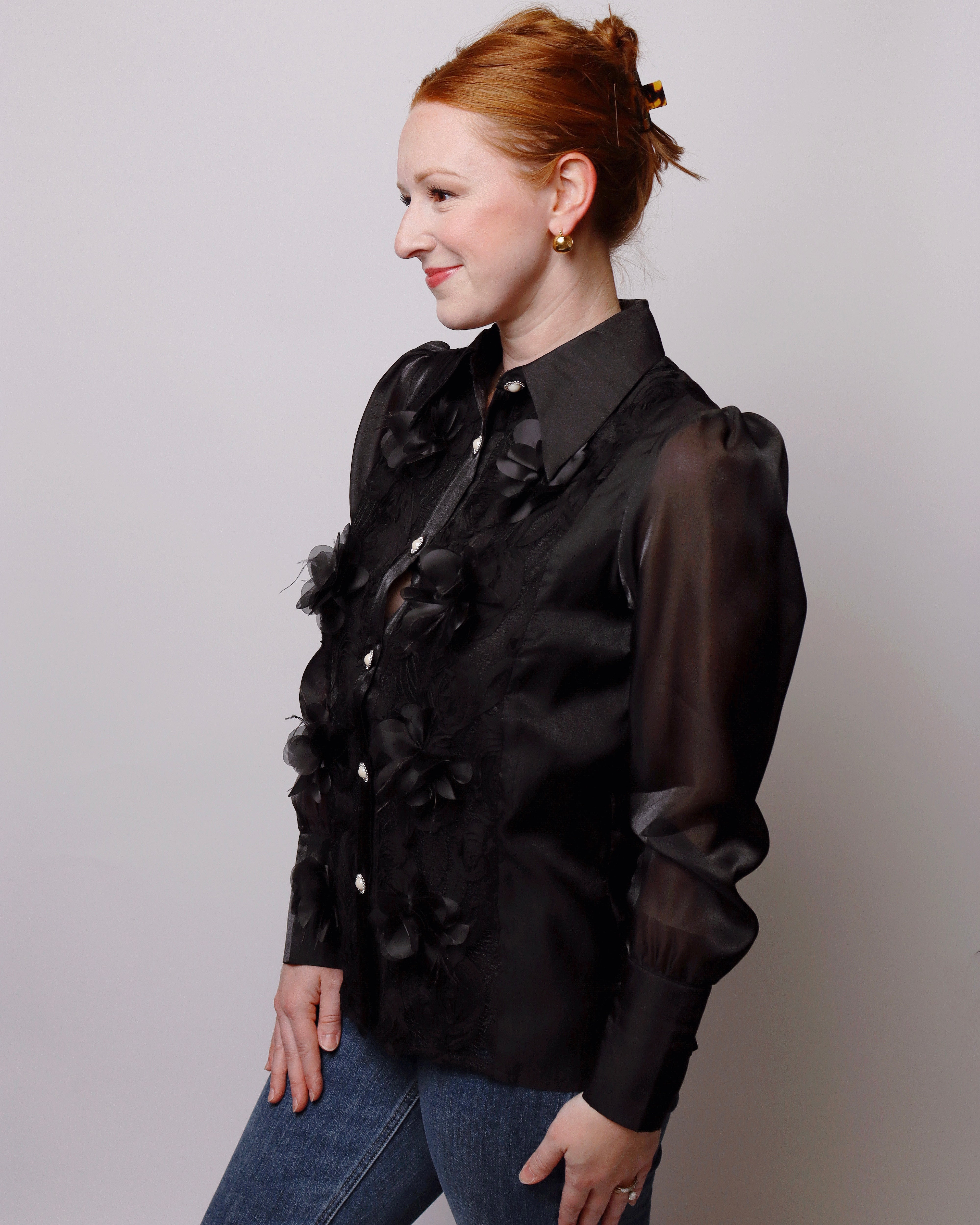 A woman with red hair tied up in a bun is wearing a black long-sleeve blouse with sheer puff sleeves and 3D floral appliques, paired with blue jeans, against a plain light gray background.