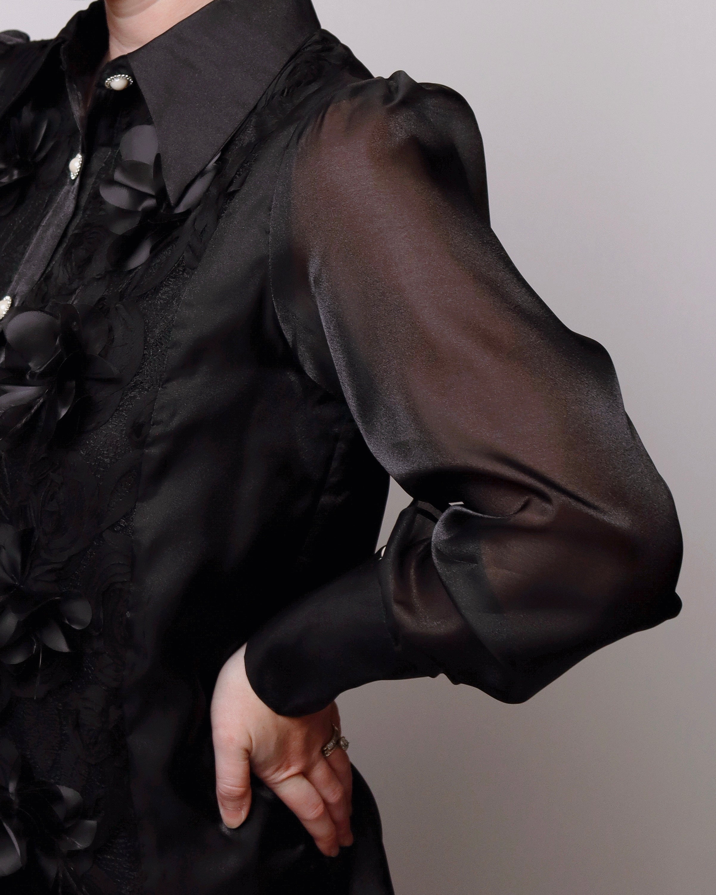 Close-up of a person wearing a black long-sleeve blouse with sheer puff sleeves, lace trim, and floral applique, against a plain light gray background.