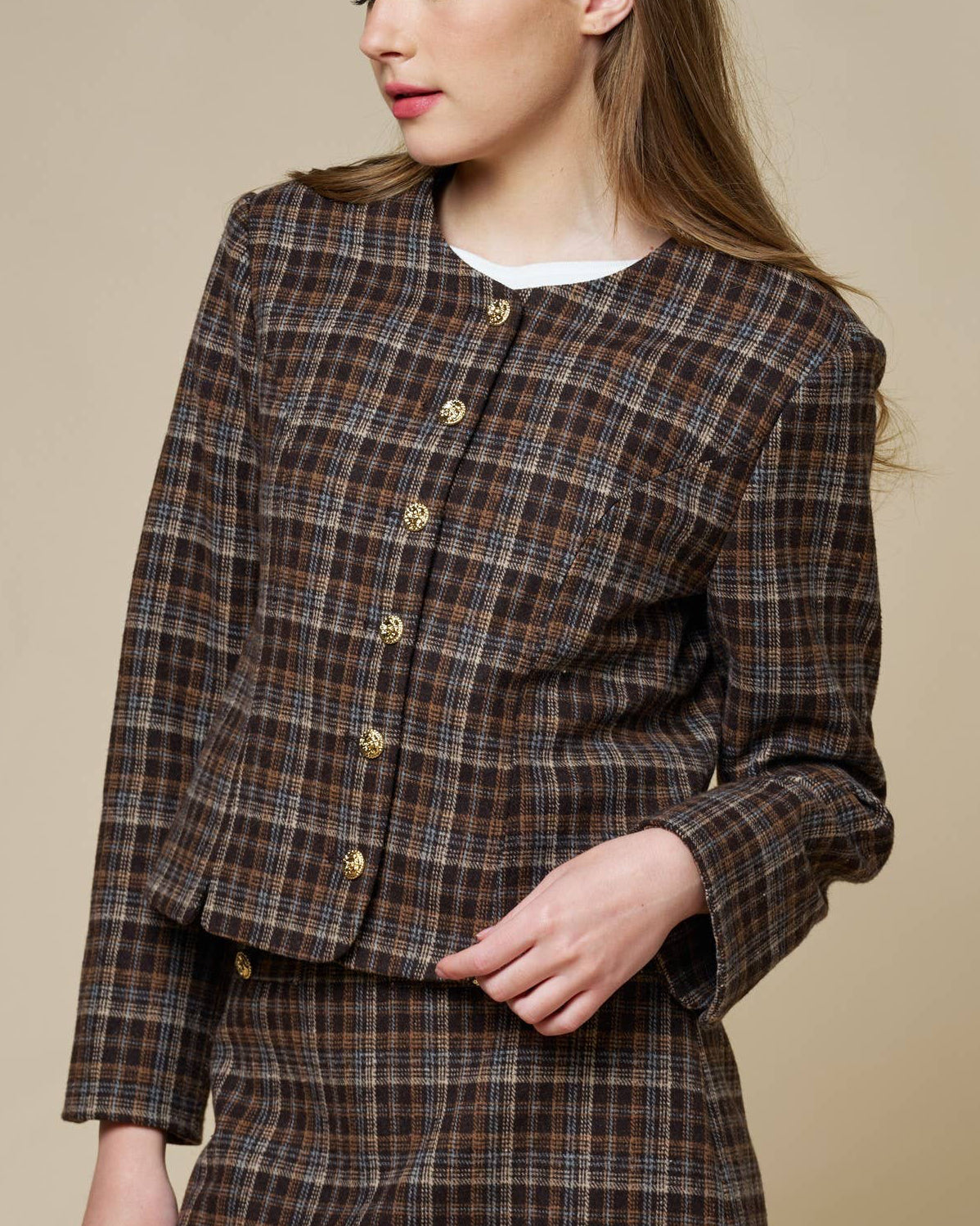 A woman modeling a brown and beige plaid tweed jacket and matching mini skirt against a solid beige studio background.