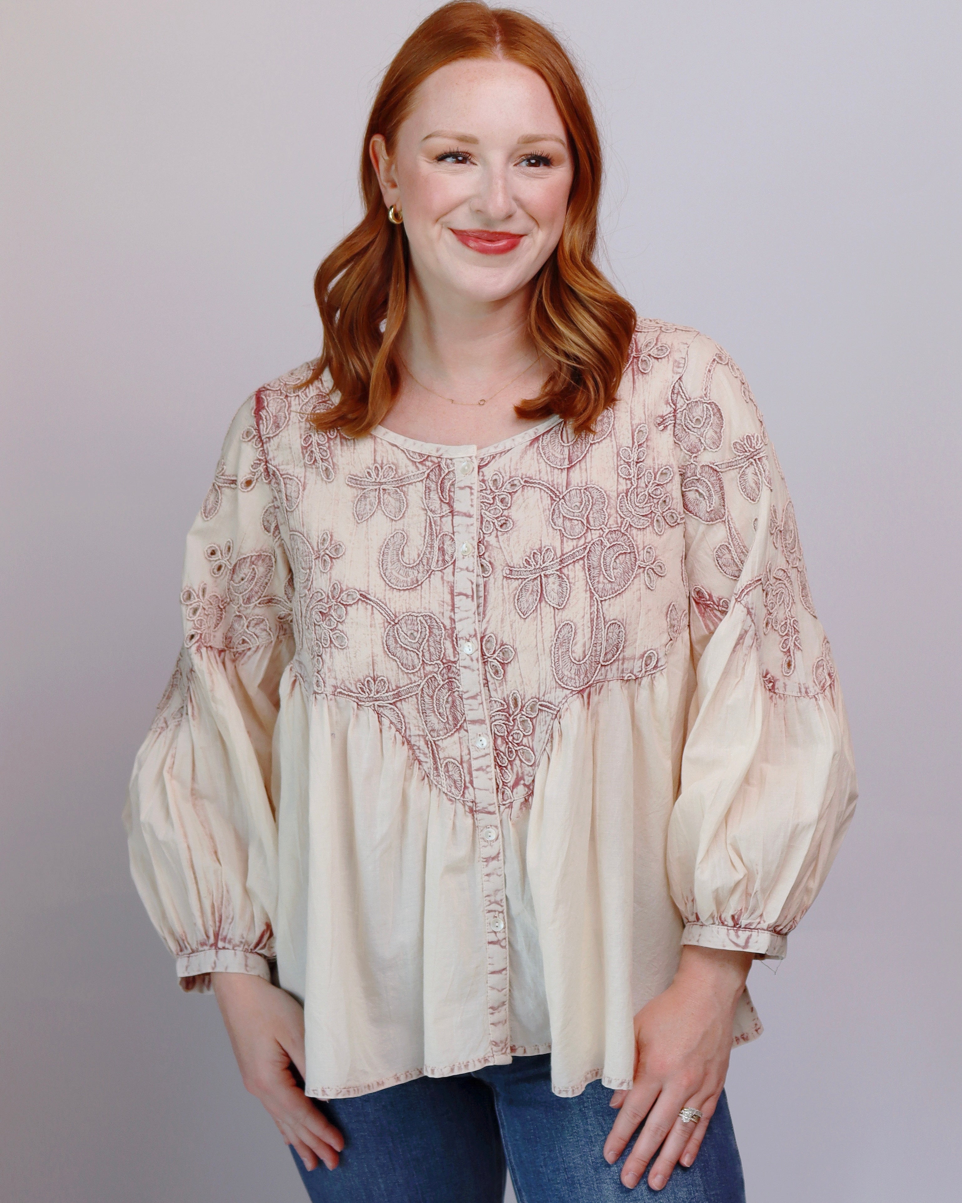A woman with red hair wearing a cream-colored long-sleeve blouse with dusty rose embroidery and blue jeans, standing against a plain light gray background.