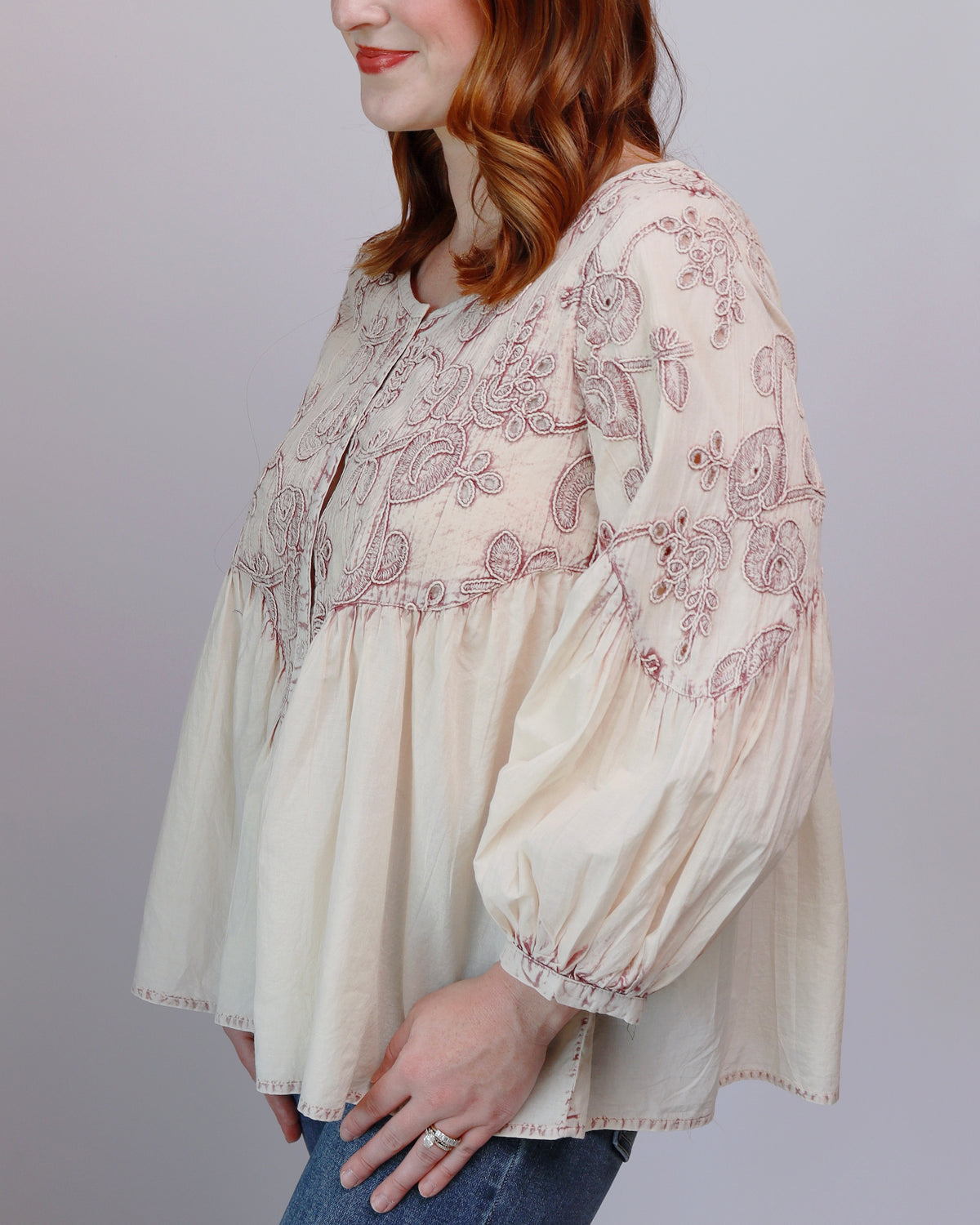 A woman wearing a cream-colored long-sleeve blouse with brown floral embroidery, paired with blue jeans, shown from the side against a plain light gray background.