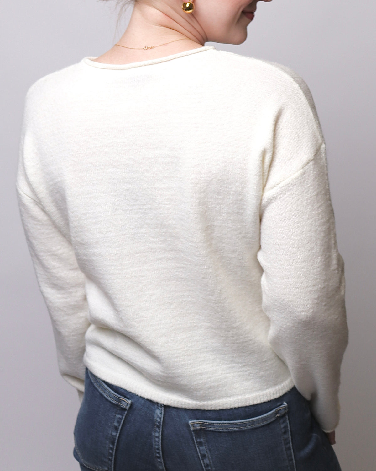 A person shown from the back wearing a soft cream knit sweater and blue jeans against a plain light gray studio background.