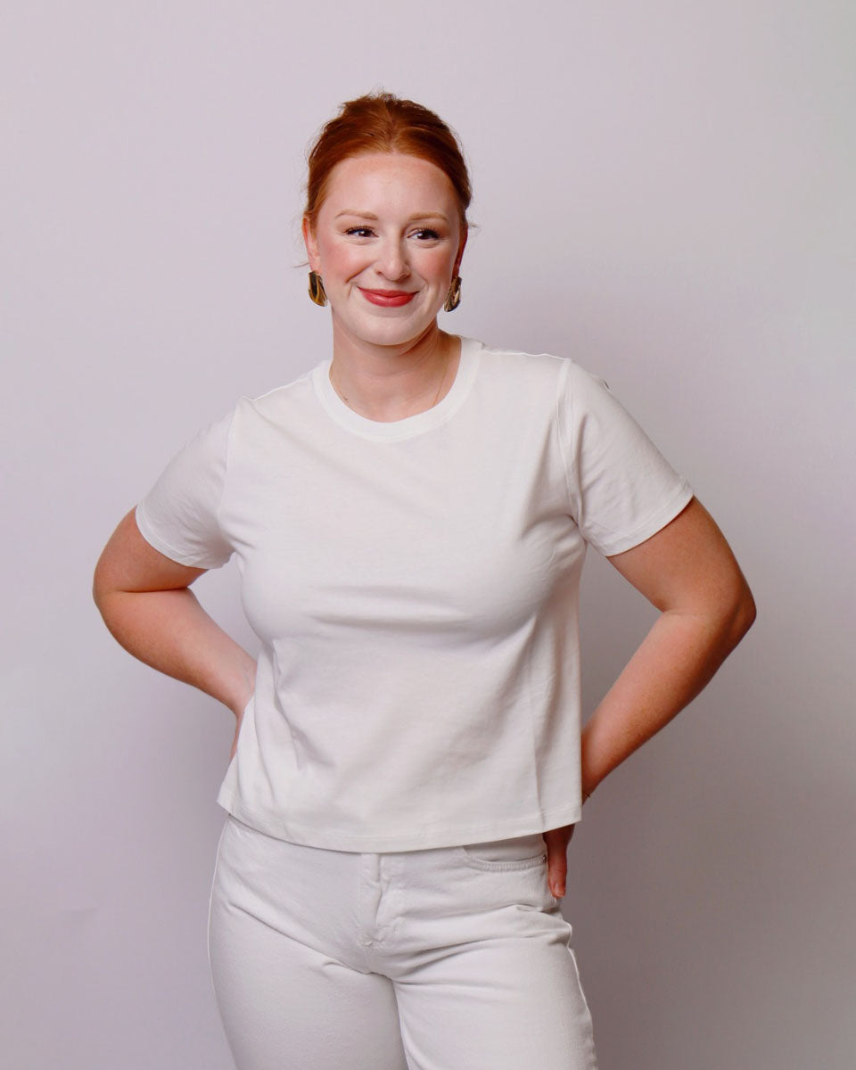 A smiling woman with red hair tied back, wearing a plain white short-sleeve crewneck t-shirt and matching white pants, standing against a light gray studio background.