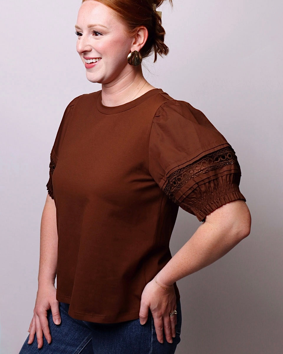A woman wearing a brown short-sleeve top with puff sleeves and lace detailing at the cuffs, paired with blue jeans, photographed against a plain light gray studio background.