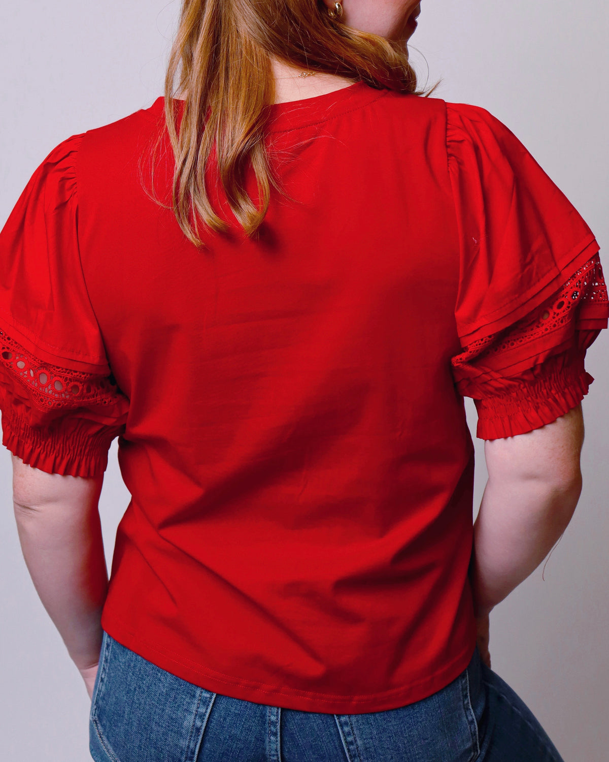 A woman viewed from the back wearing a bright red short-sleeve blouse with lace-trimmed ruffled sleeves and blue jeans, against a plain light gray background.