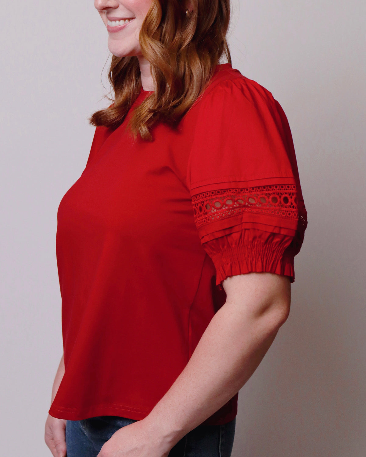 A woman wearing a bright red short-sleeve top with eyelet lace detailing on the sleeve cuffs, paired with blue jeans, shown against a plain light gray background.