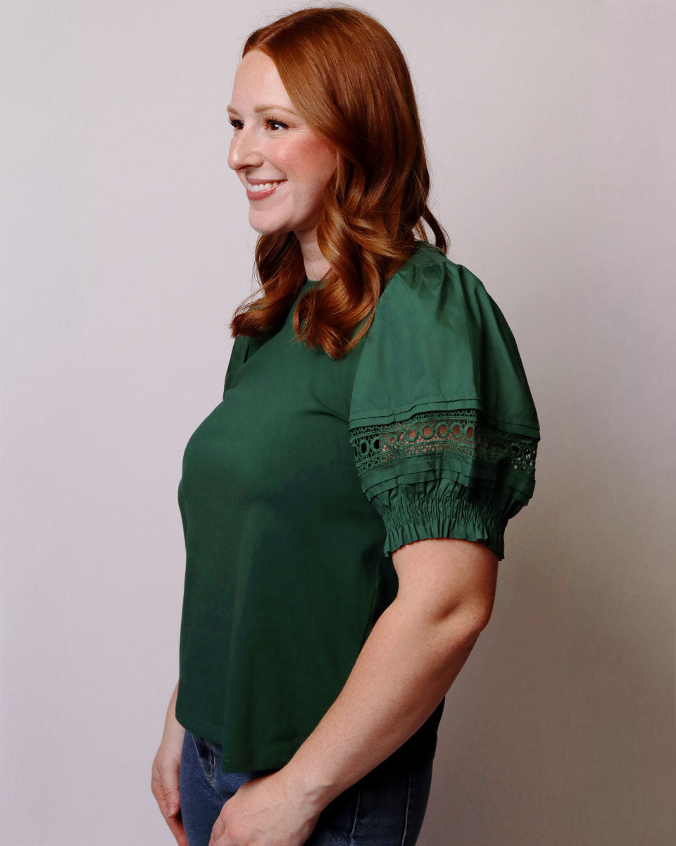 A woman with red hair wearing a dark green short-sleeve blouse with puff sleeves and embroidered details on the upper sleeve, paired with blue jeans, standing against a plain light gray background.