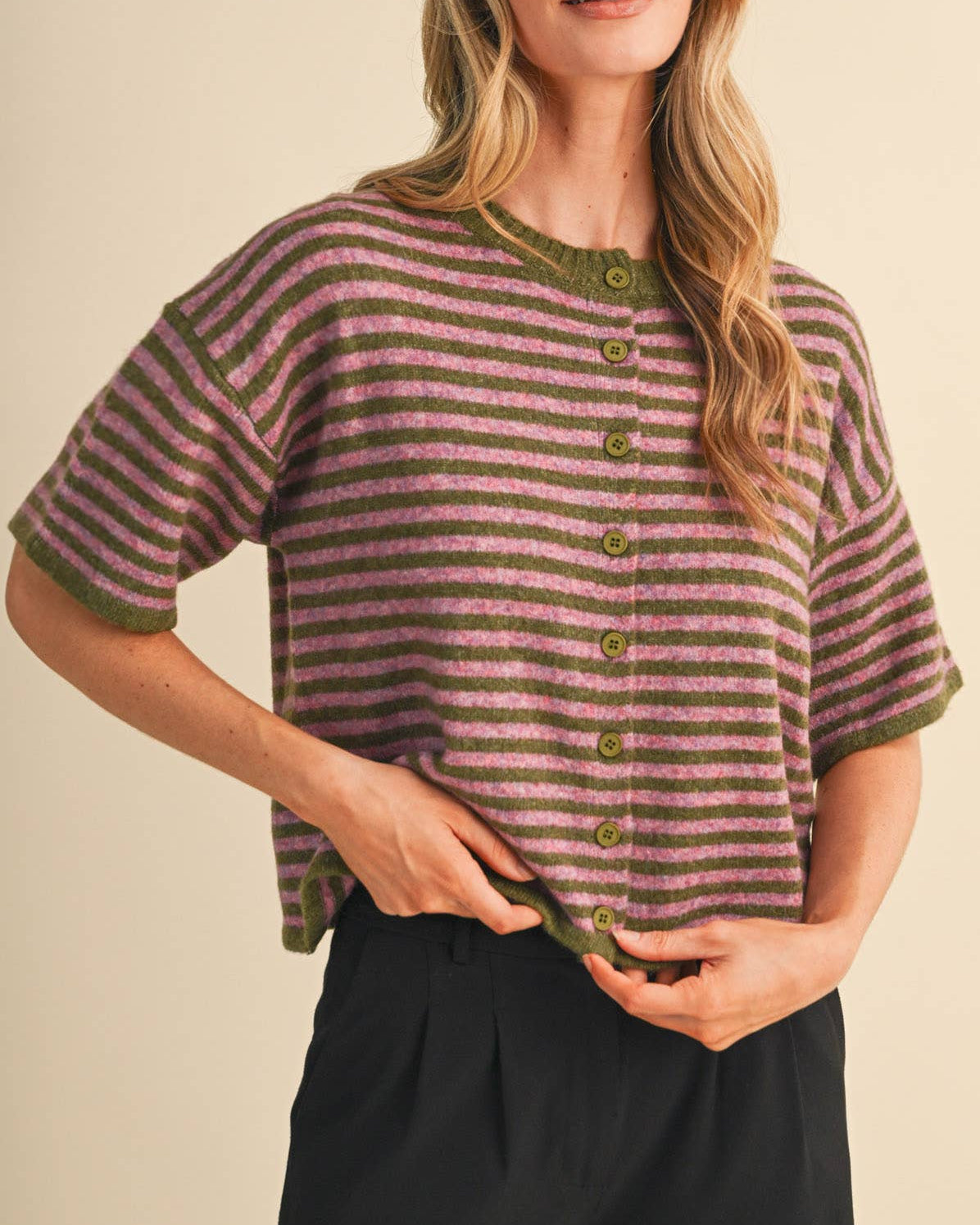 A woman modeling a short-sleeve knit top with horizontal stripes in orchid and moss green, featuring a button-front closure and matching moss trim, on a neutral background.