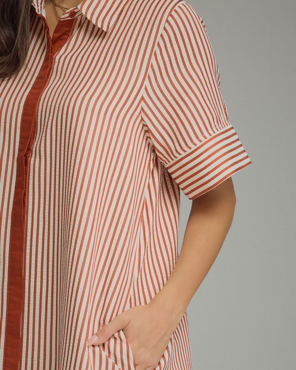 A close-up of a woman wearing a short-sleeve collared shirt dress with vertical rust-orange and off-white stripes, featuring a button placket and side pocket.