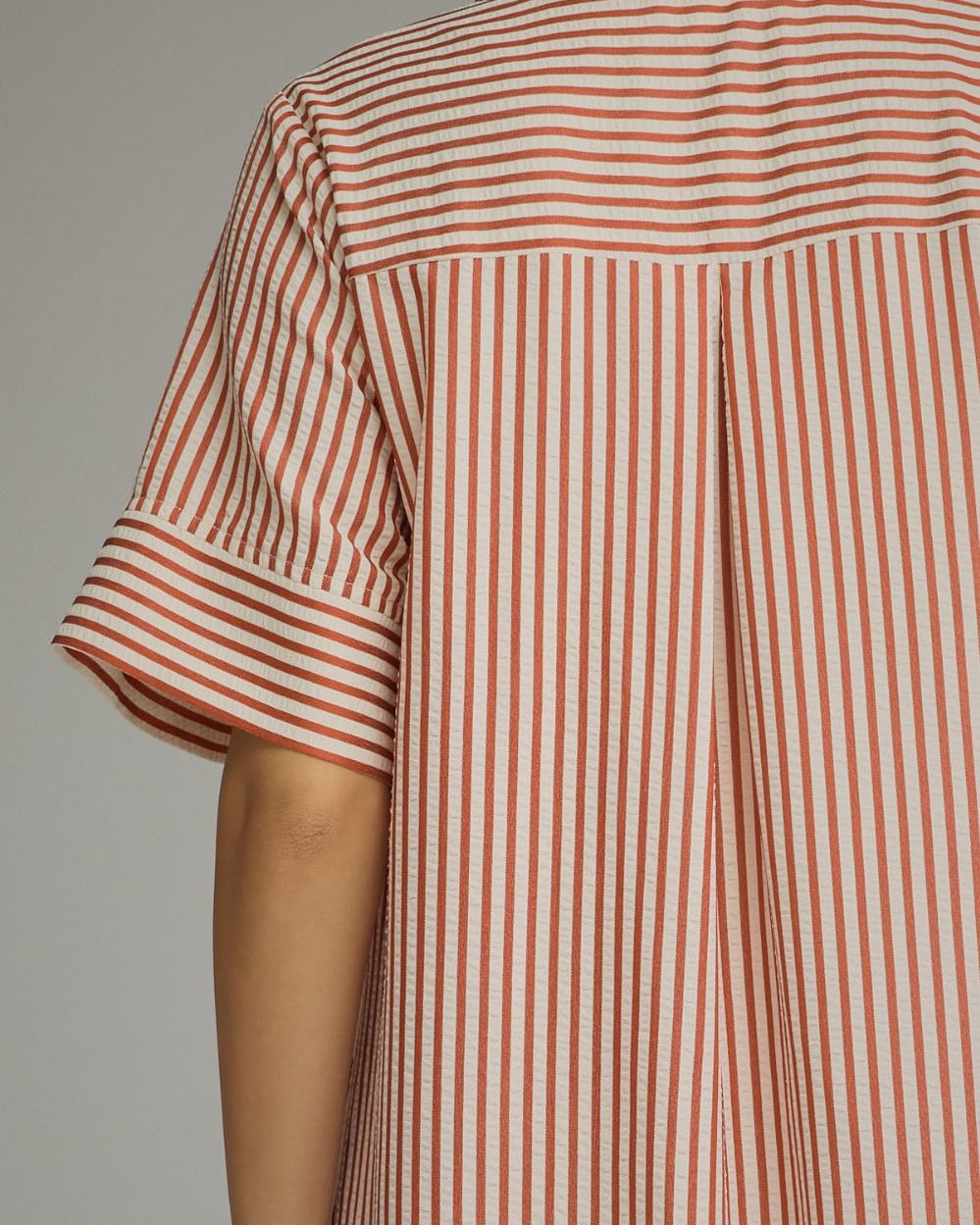 Close-up of a person wearing a short-sleeve collared shirt with vertical red and beige stripes, shown from the back against a plain light gray background.