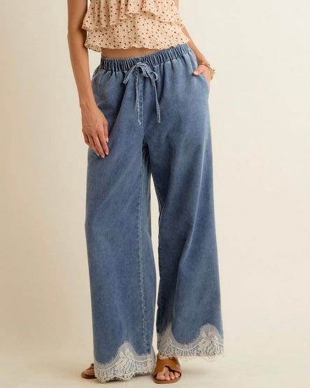 A person wearing wide-leg blue denim pants with a drawstring waist and white lace trim at the hem, paired with a beige polka-dot ruffled top and brown sandals, against a plain cream background.