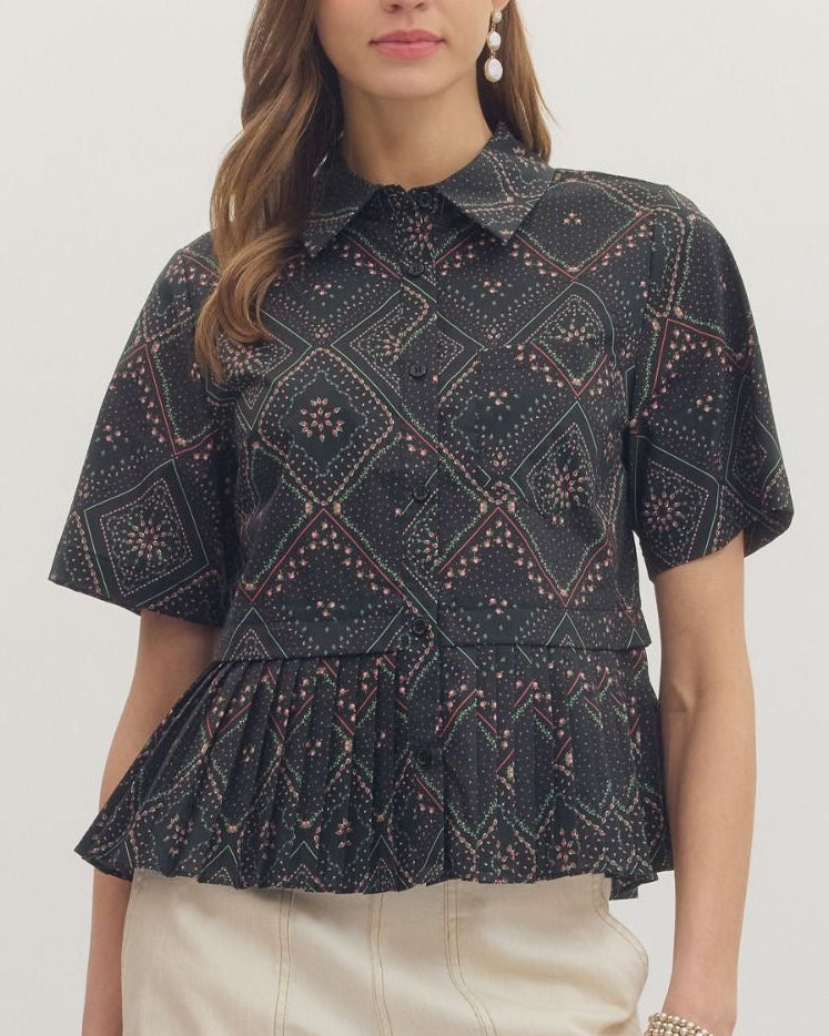 A woman modeling a short-sleeve collared blouse with a black base and intricate geometric and floral patterns in beige and muted pink on a neutral background