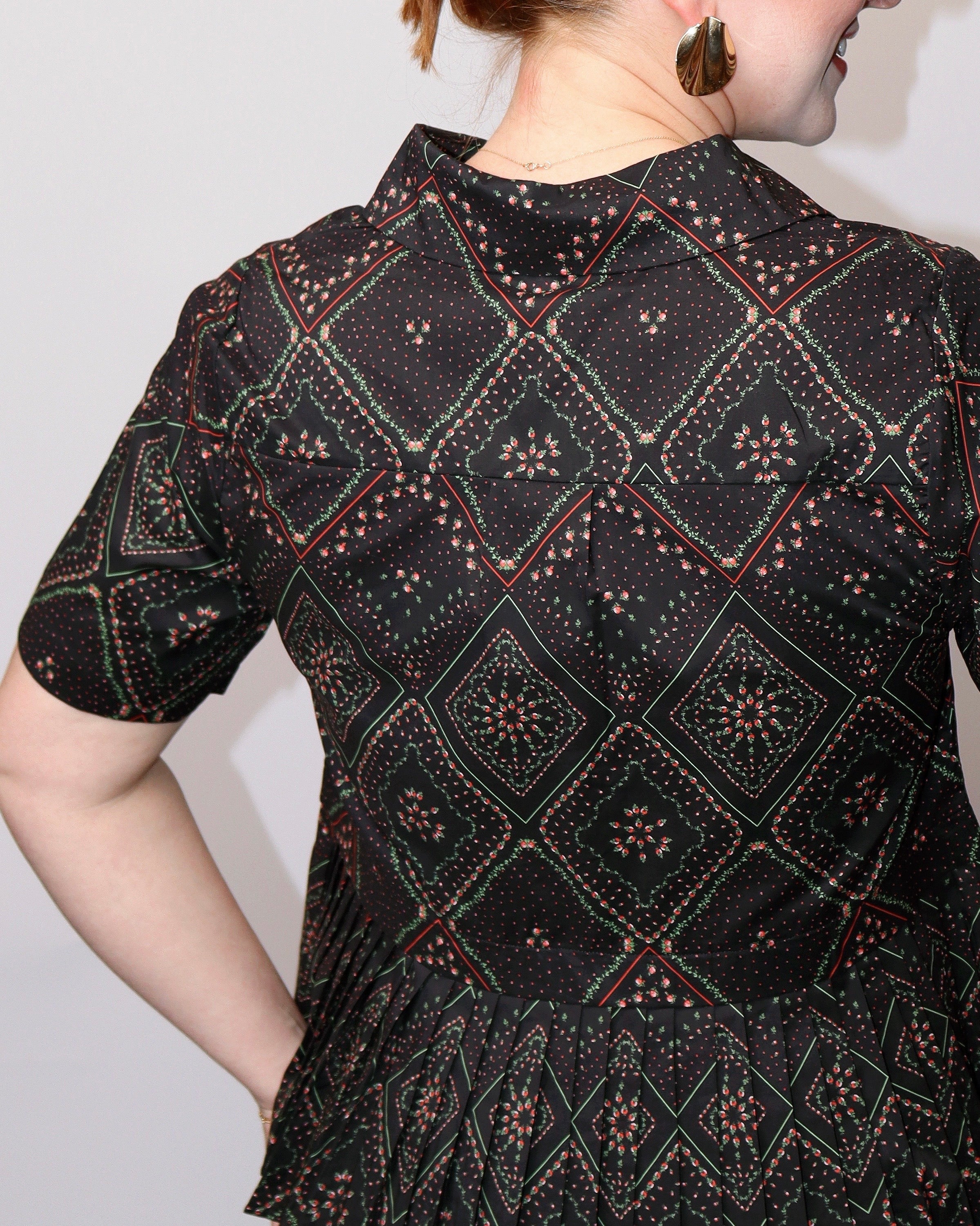 A close-up of a person wearing a short-sleeve black dress with a geometric diamond pattern in red, green, and beige, photographed against a plain light background.