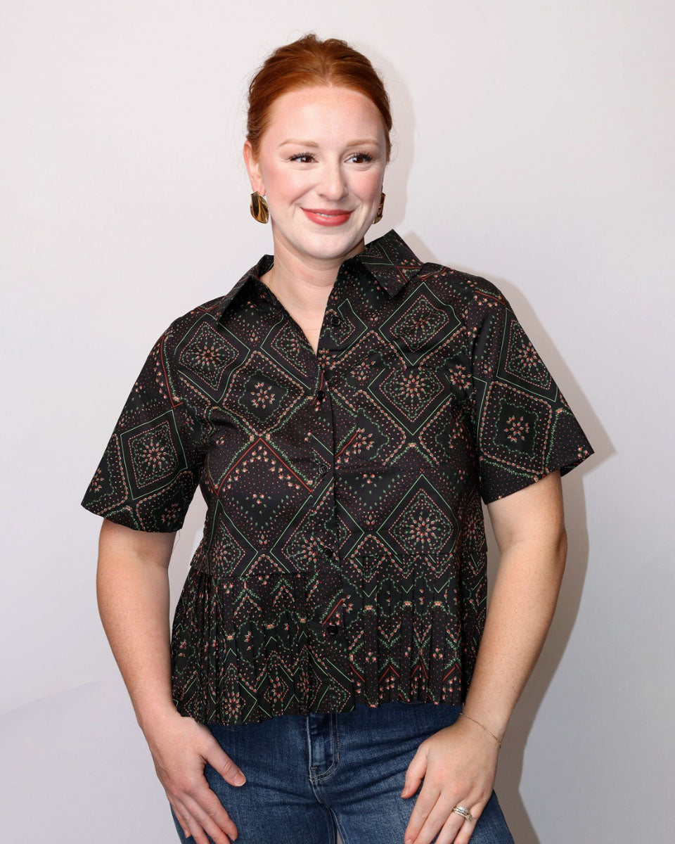 A woman with red hair wearing a black short-sleeve button-up shirt with a brown and beige geometric pattern, paired with blue jeans, standing against a plain light gray background.
