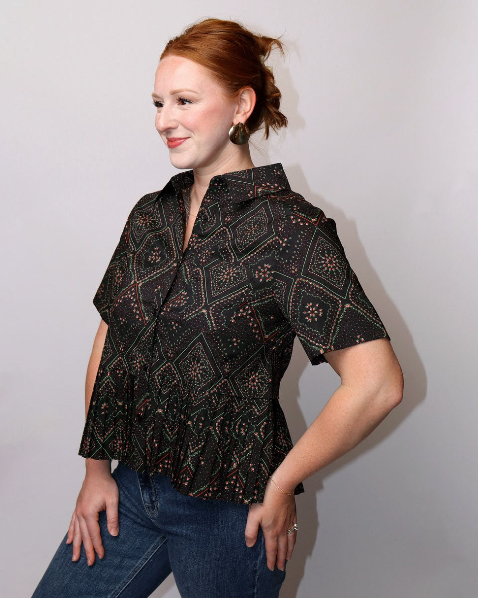 A woman with red hair tied back wears a short-sleeve button-up blouse with a black base and a brown geometric pattern, paired with blue jeans, posing against a light gray background.
