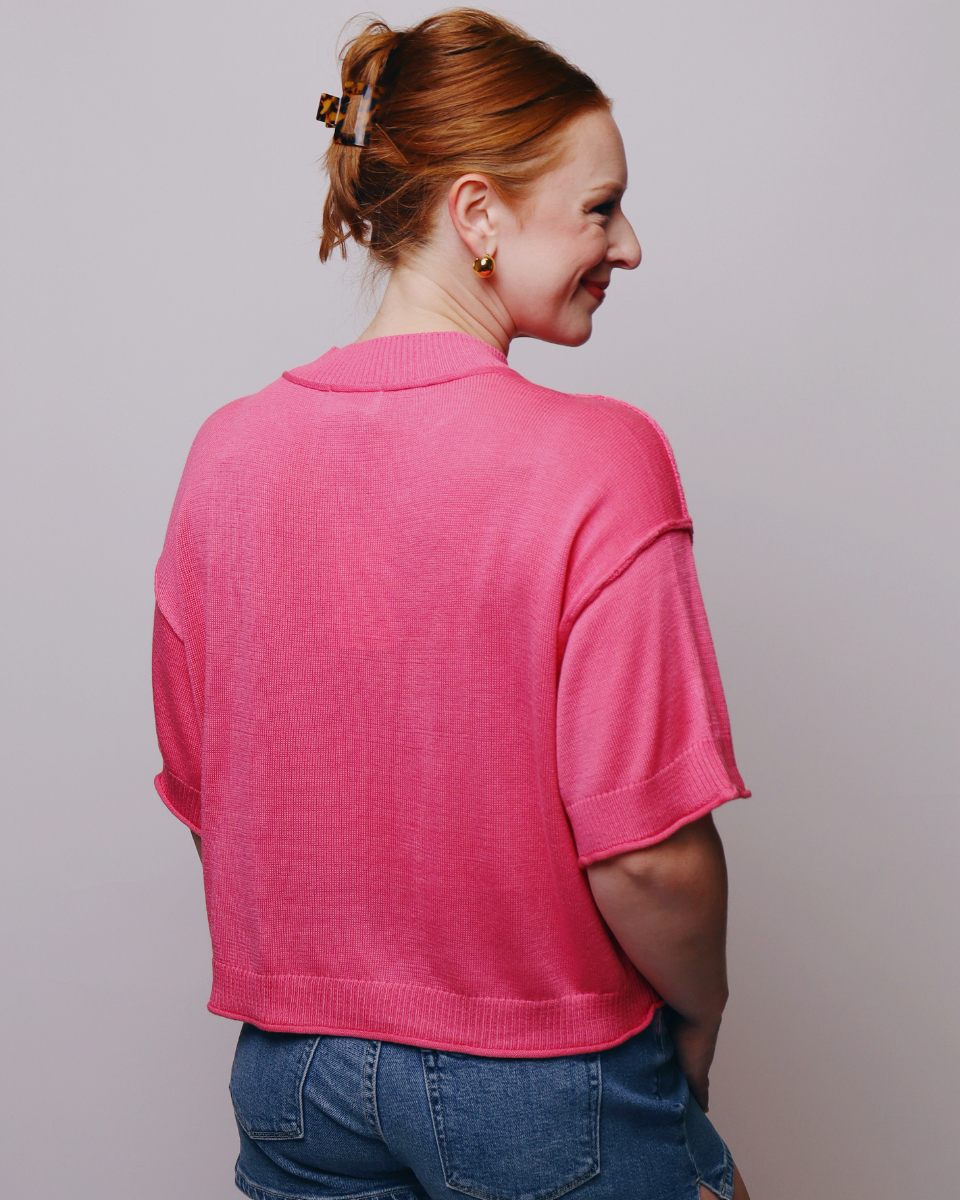 A woman with red hair tied up in a bun, viewed from behind, wearing a bright pink short-sleeve knit top and blue jeans against a plain light gray background.