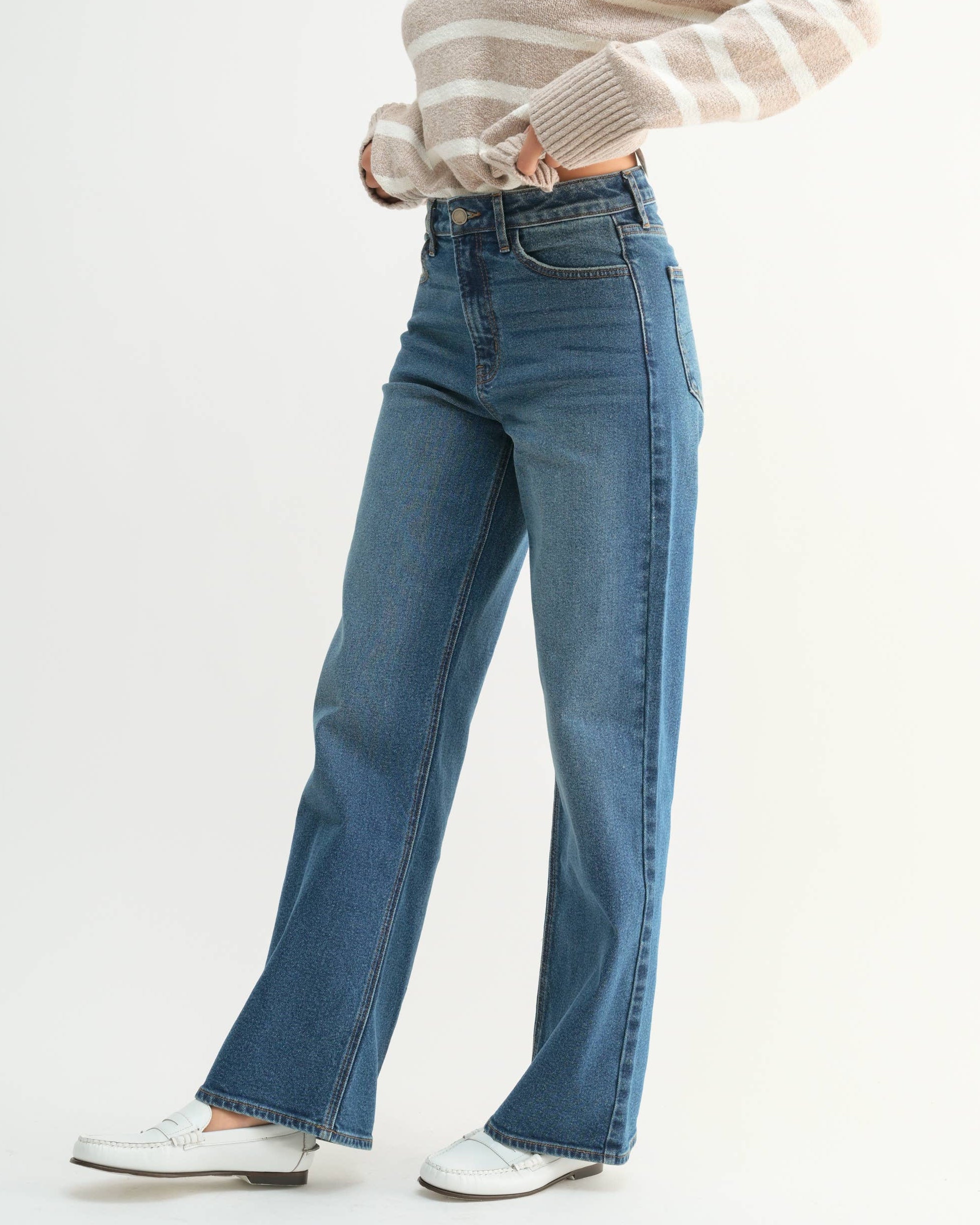 A woman modeling a beige and white striped knit polo sweater paired with high-waisted, wide-leg blue jeans and white sneakers, set against a plain white studio background.