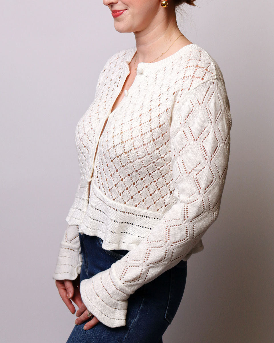 A woman wearing a cream-colored, open-knit cropped cardigan with diamond-patterned sleeves, paired with dark blue jeans, shown from a side profile against a neutral gray background.