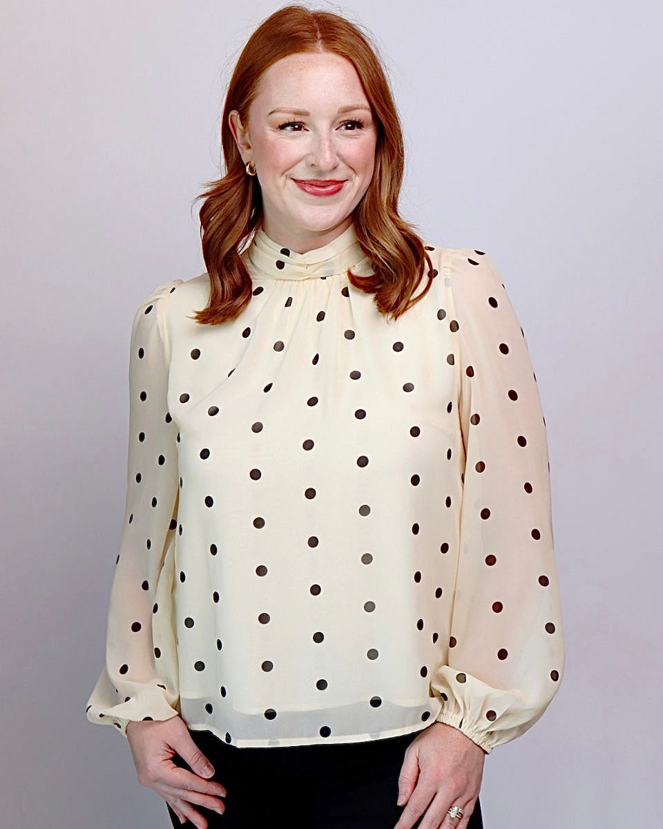 A woman with red hair wearing a cream-colored long-sleeve blouse with black polka dots and a high gathered neckline, paired with black pants, against a light gray studio background.