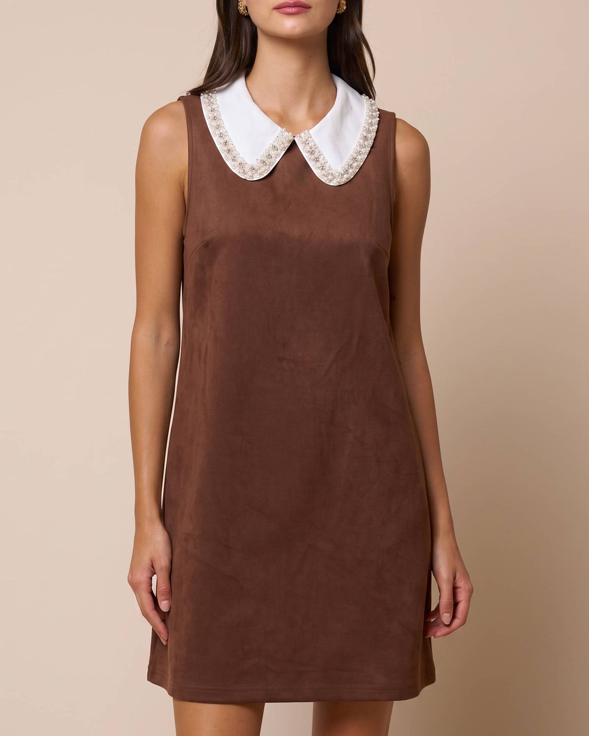 A woman modeling a sleeveless brown dress with a contrasting white Peter Pan collar trimmed with delicate gold lace, set against a solid beige background. Isabella Rhinestone Contrast Collar Faux Suede Mini Shift Dress | Blackbird Boutique