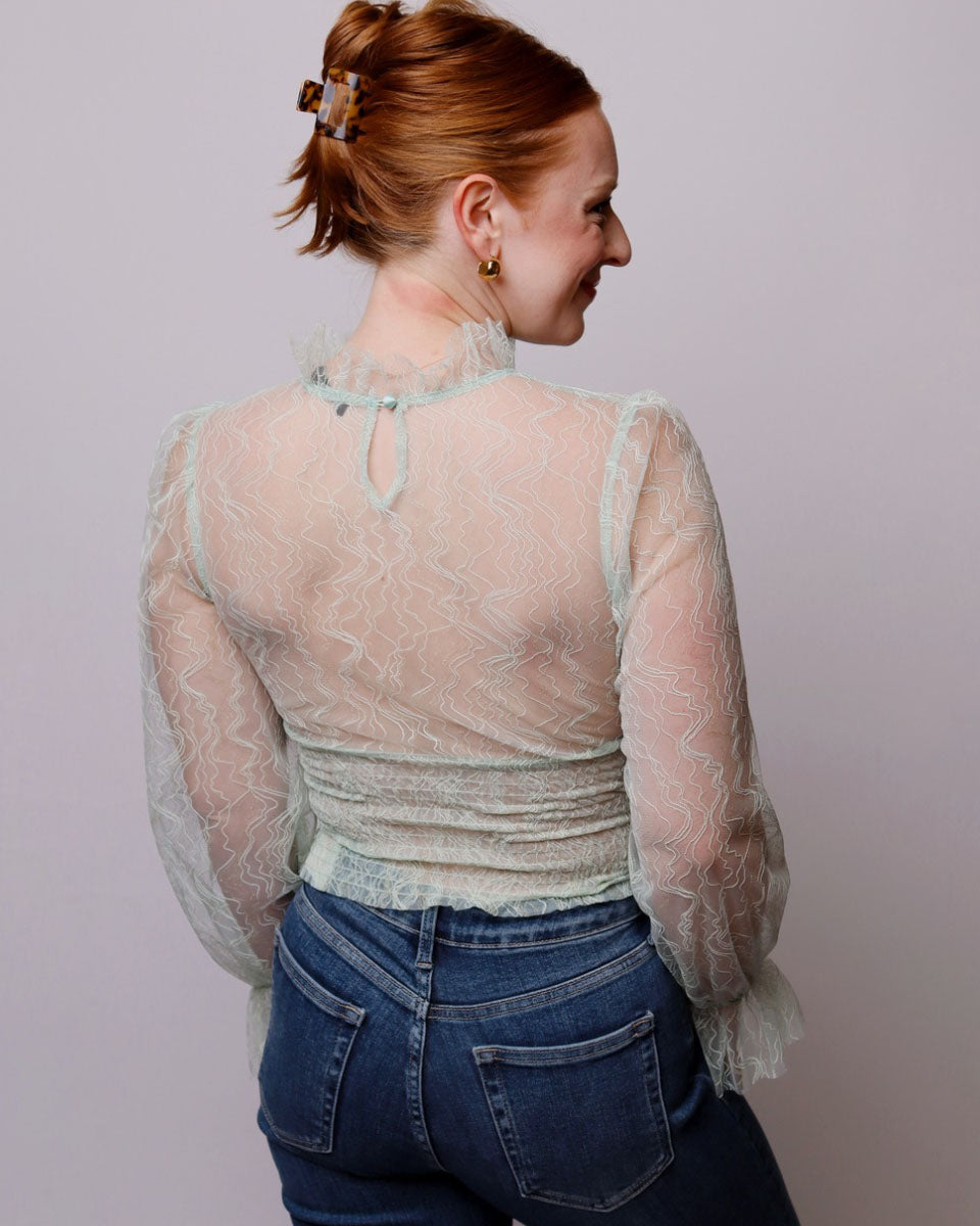 Woman wearing a sheer lace top over a patterned shirt with blue jeans against a plain background