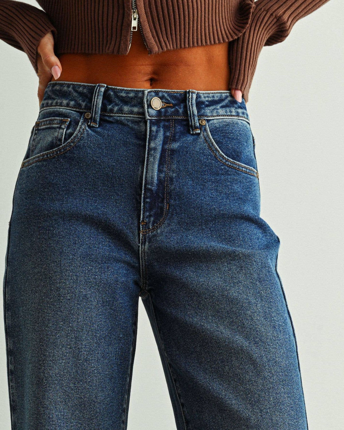 A person wearing dark blue wide-leg denim jeans, a brown ribbed crop top, and brown heeled sandals, standing against a light gray wall and green floor.