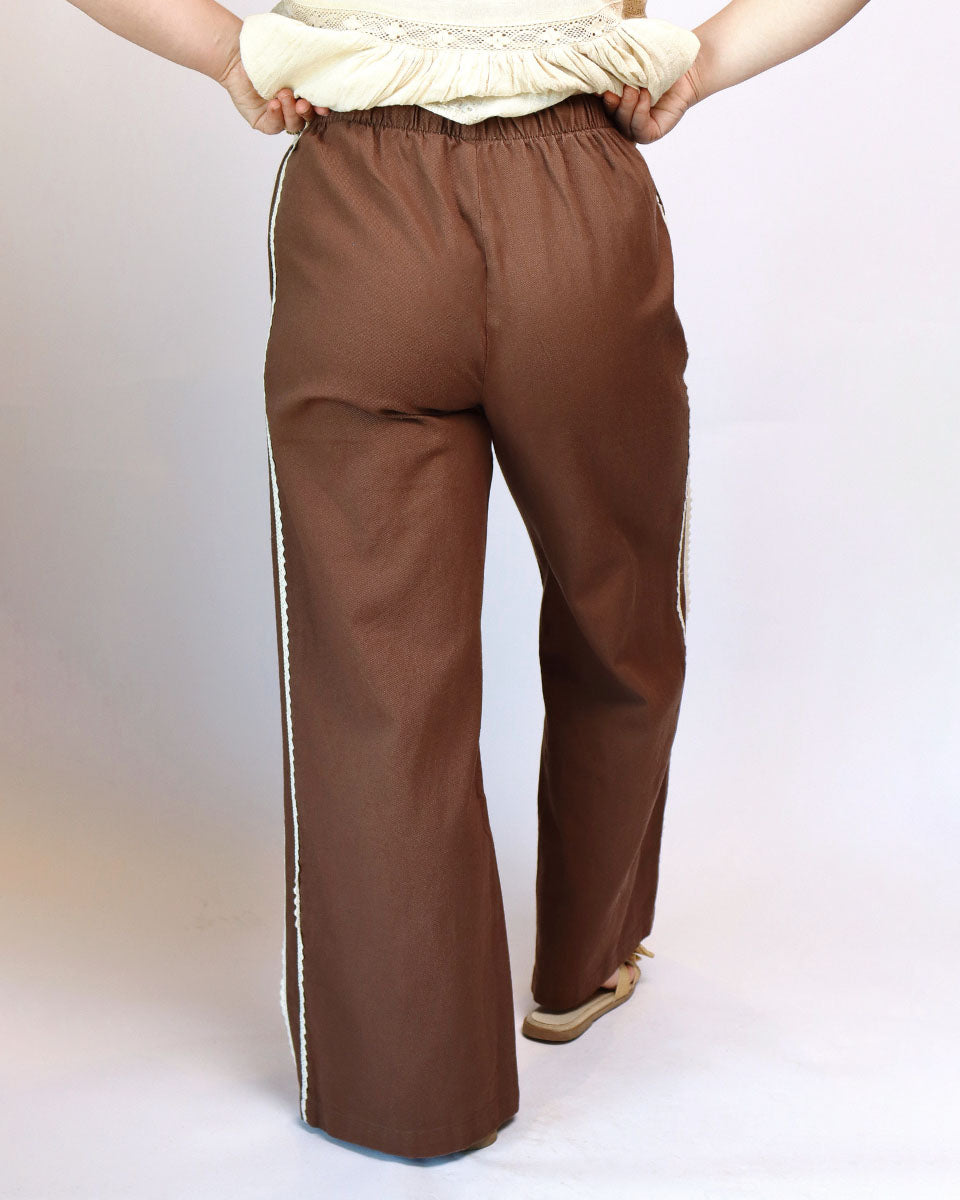 A person shown from the back wearing brown wide-leg pants with white side stripes, holding the waistband of a cream top.
