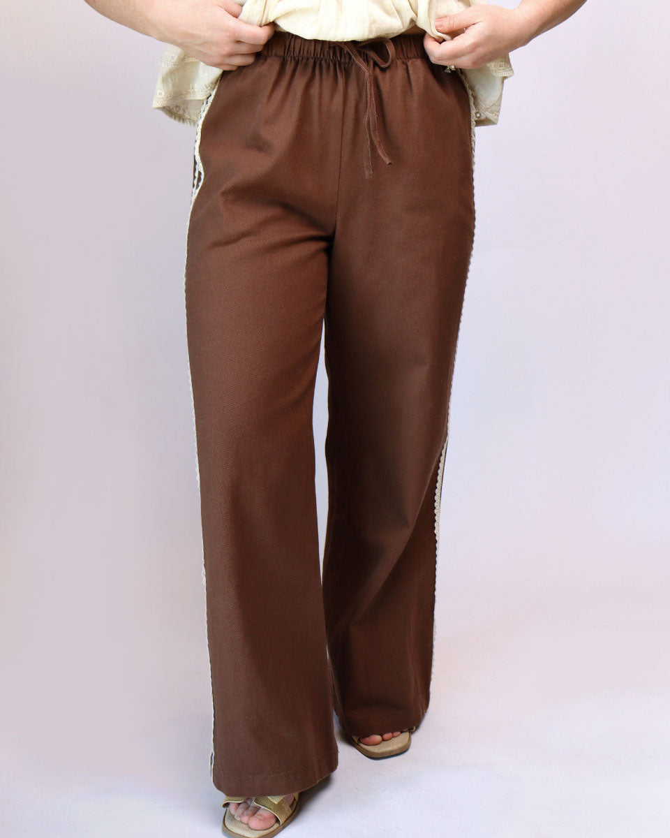 A person wearing brown wide-leg drawstring pants with light-colored side stripes, paired with a cream top and sandals, shown against a plain light background.