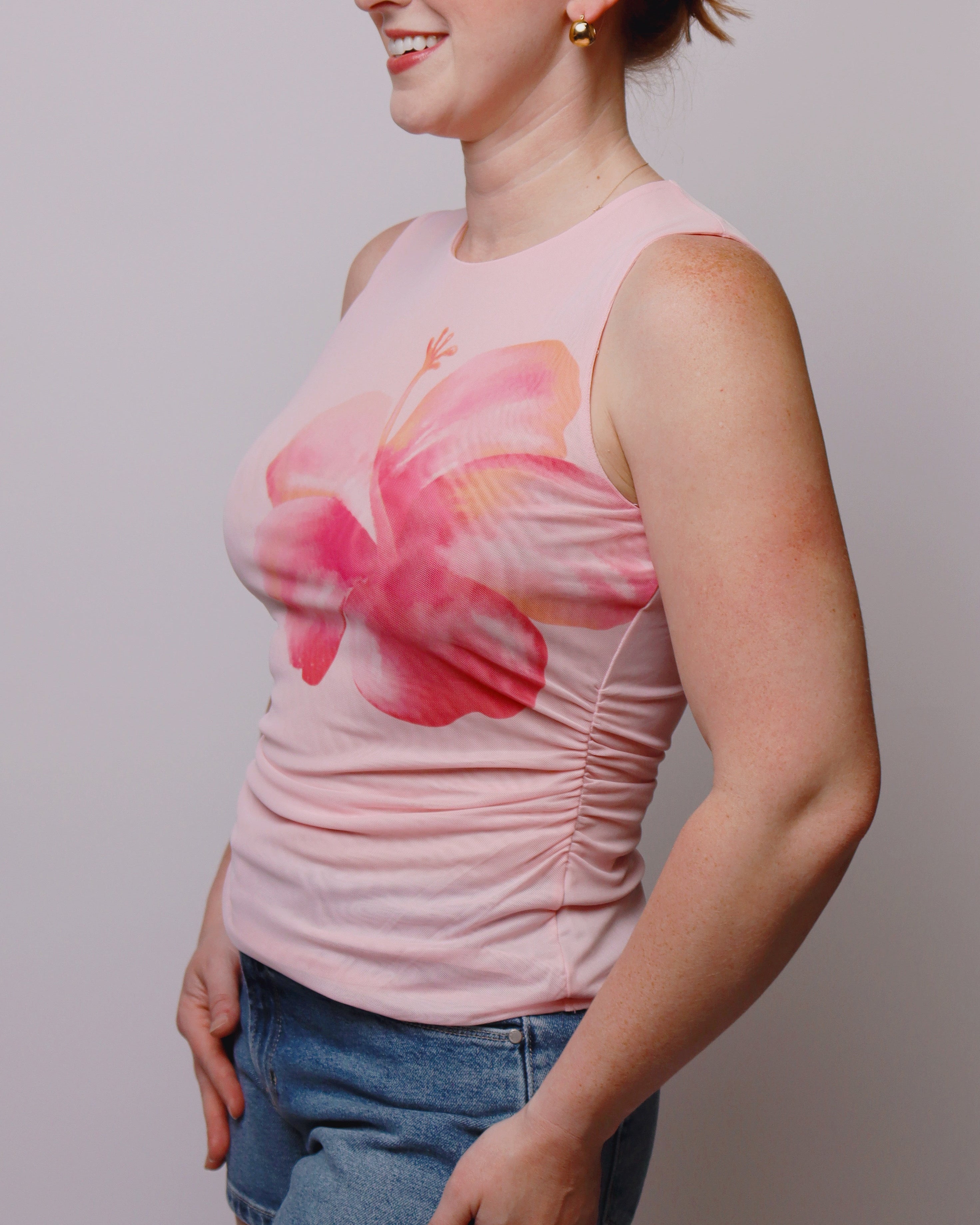Woman wearing a pink sleeveless top with a floral design and denim shorts against a plain background