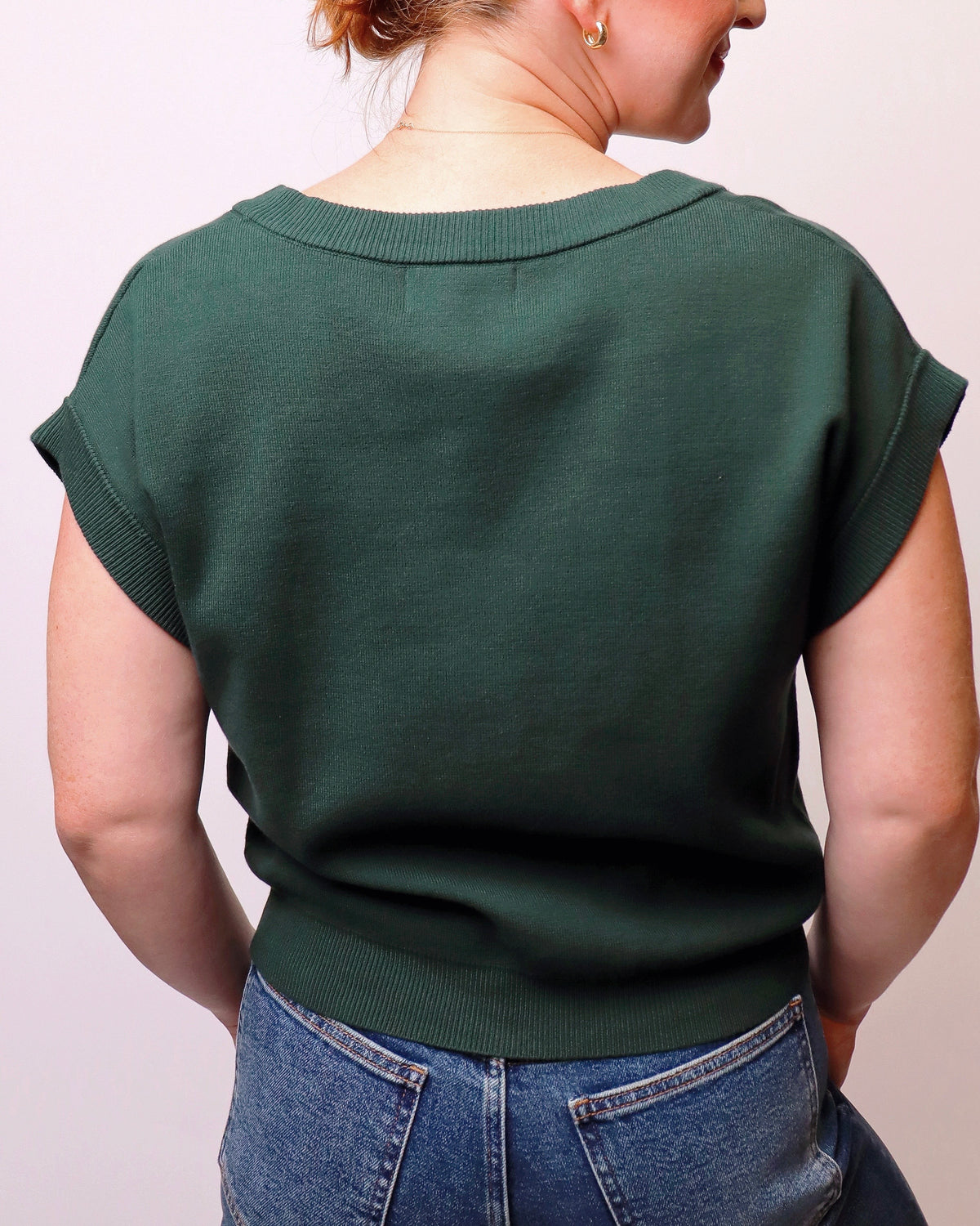A woman shown from the back wearing a dark forest green short-sleeve knit top and blue jeans against a plain light background.
