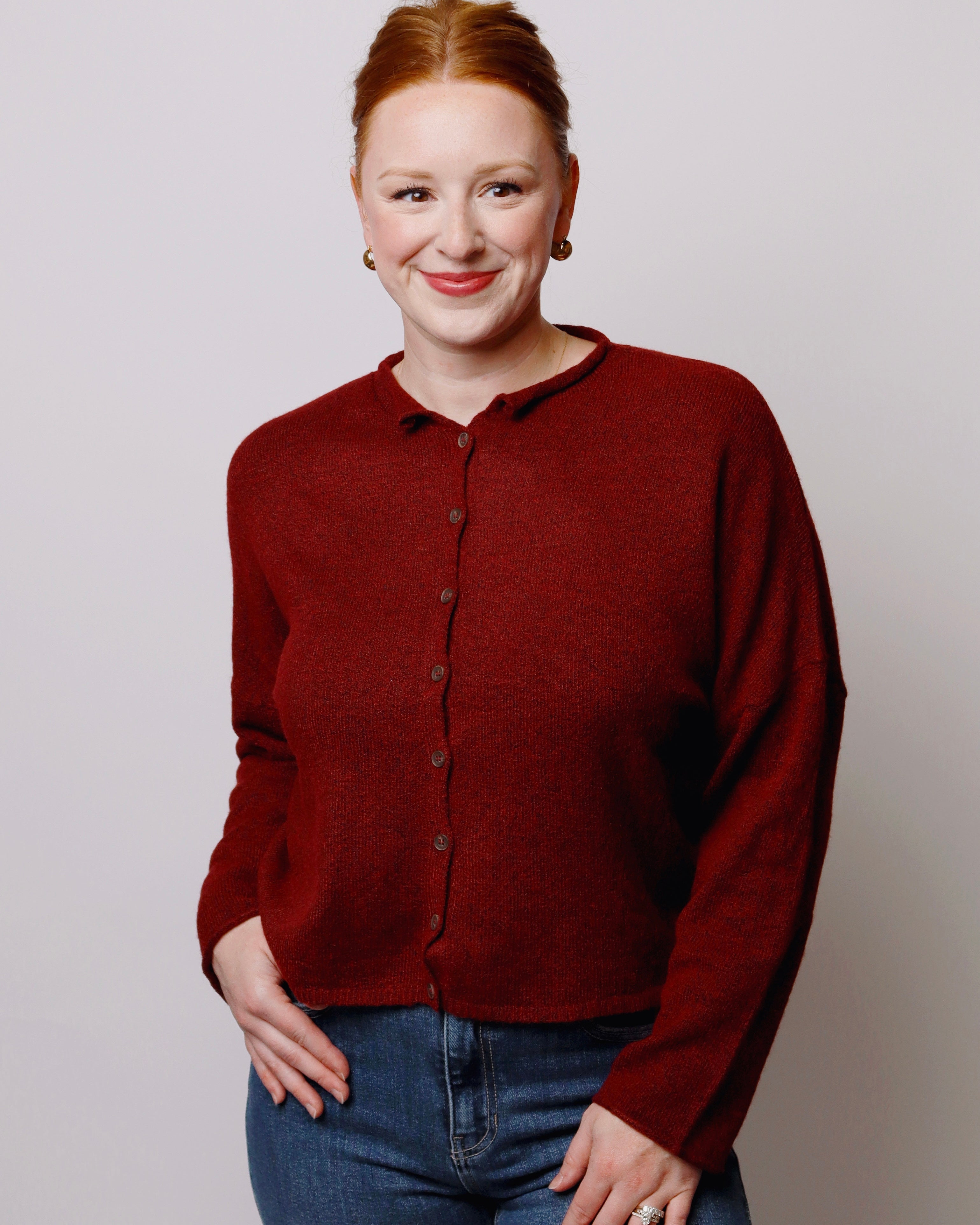 A woman with red hair tied back, wearing a dark red button-up knit cardigan and blue jeans, standing against a light gray studio background.