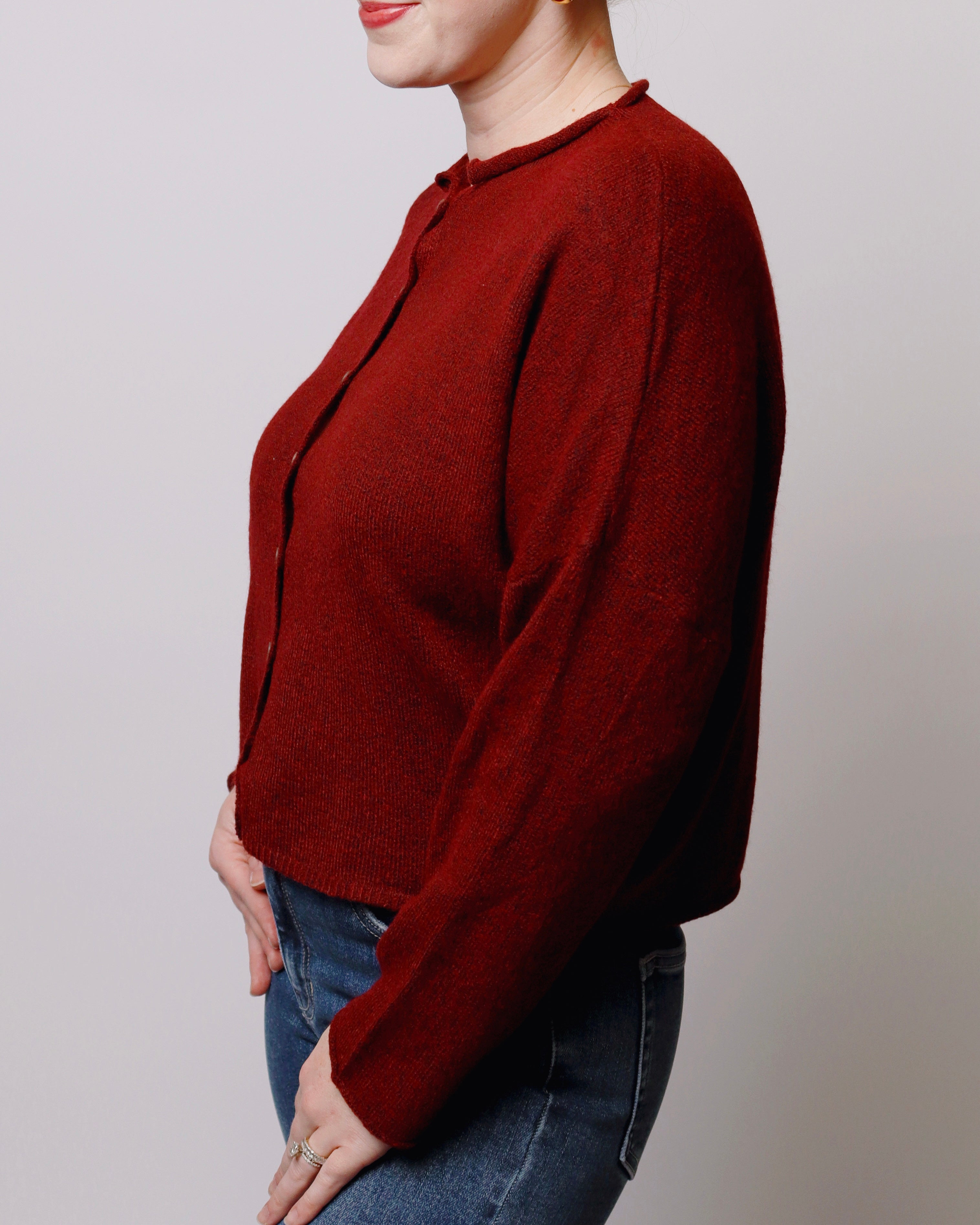 A woman wearing a dark red knit cardigan sweater with long sleeves and a scalloped neckline, paired with blue jeans, shown from a side profile against a light gray background.