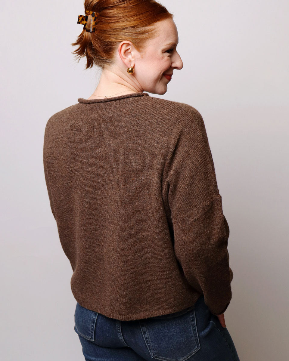 A woman with red hair tied back in a bun, viewed from behind, wearing a brown knit sweater and blue jeans against a plain light gray background.