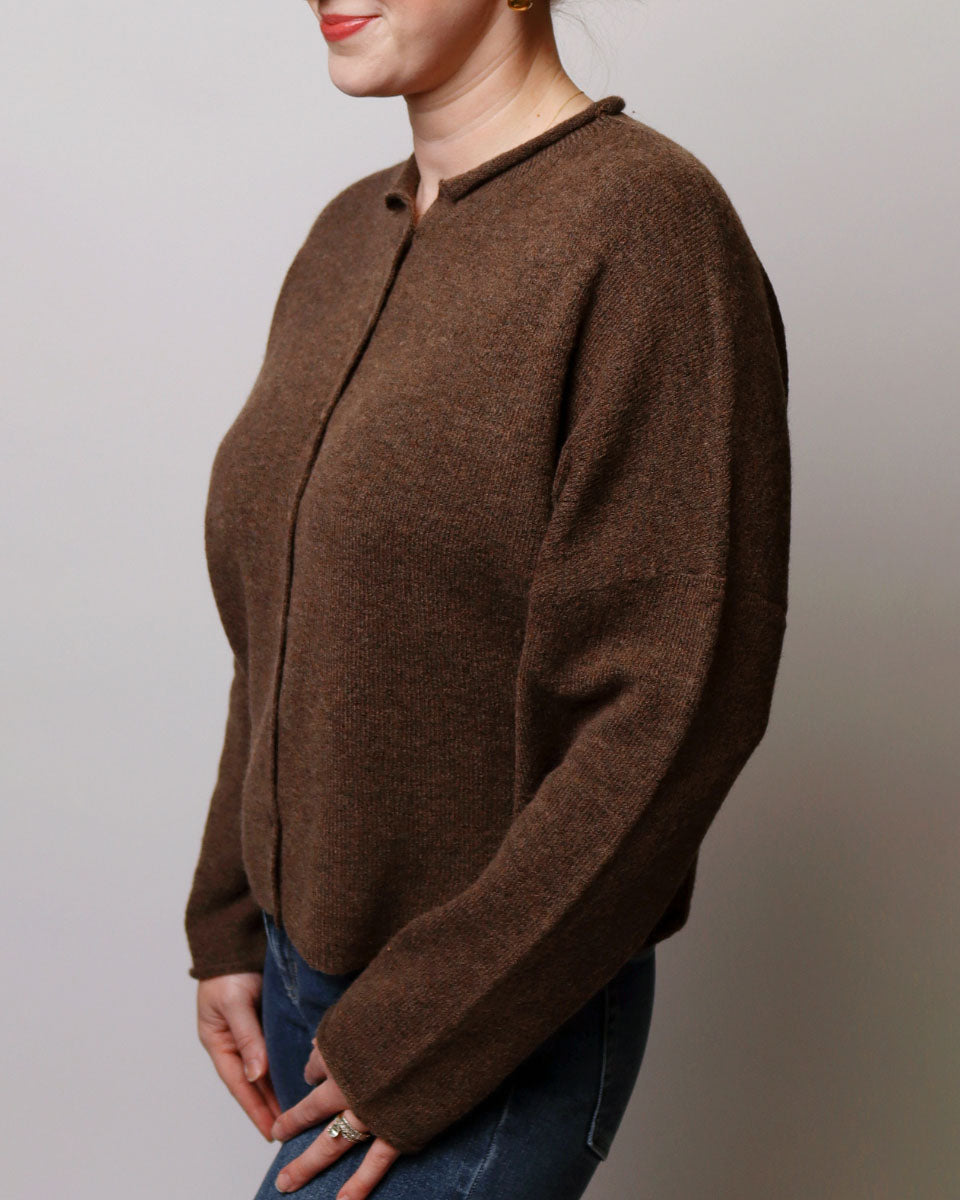 A woman wearing a brown knit cardigan sweater with a buttoned front and long sleeves, paired with blue jeans, shown from the side against a plain light gray background.