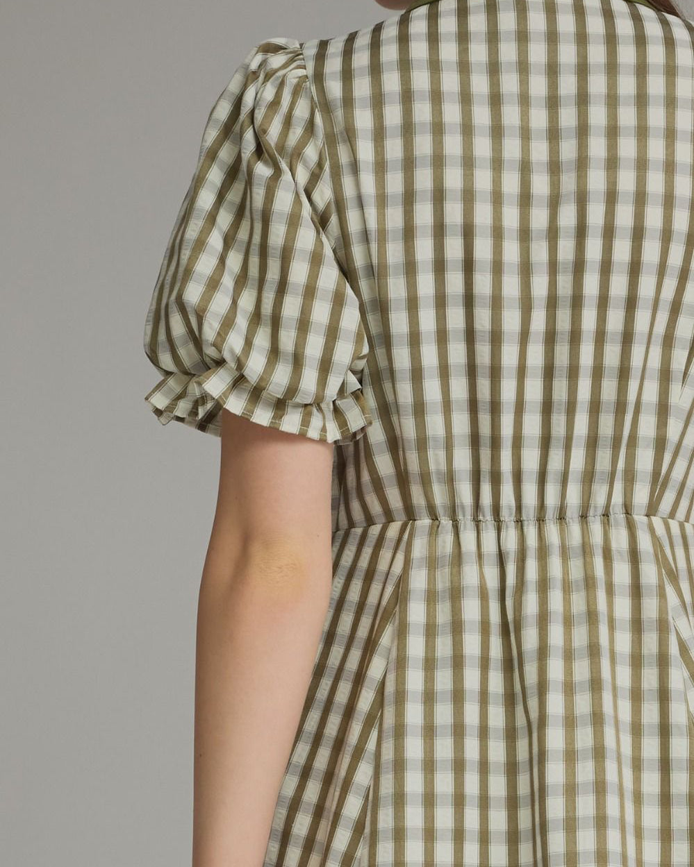 Close-up back view of a woman wearing a short-sleeve dress with an olive green and beige gingham pattern, featuring a solid olive green collar and gathered waist.