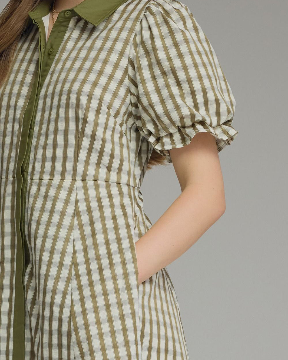 A close-up of a model wearing a short-sleeve gingham dress in olive green and cream, featuring a solid olive green collar and front placket.