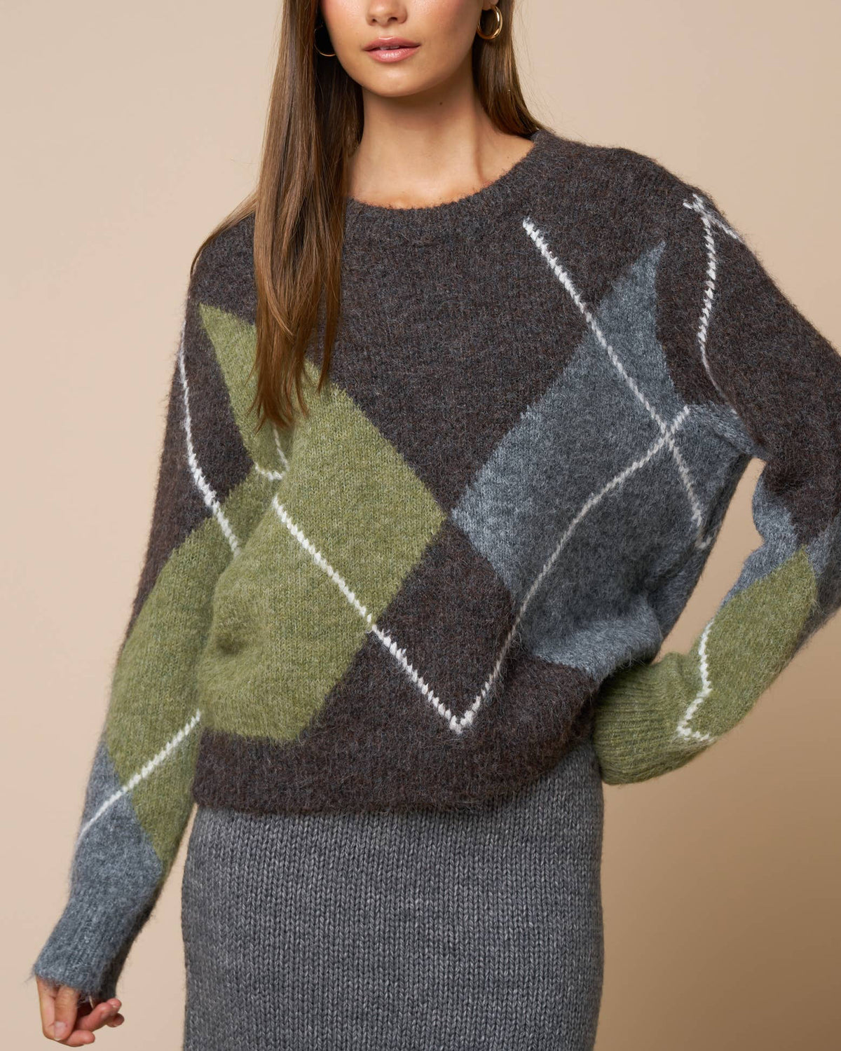 A woman wearing a fuzzy knit sweater with an argyle pattern in dark gray, olive green, slate blue, and white, paired with a gray skirt, against a solid beige background.