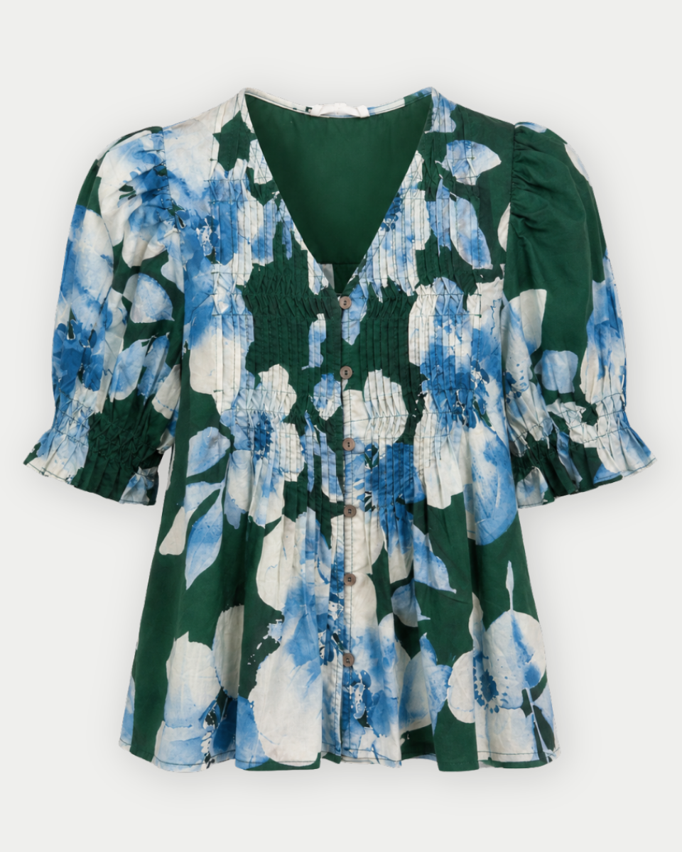 A women's short-sleeve blouse with a smocked bodice, V-neckline, and ruffled sleeves, featuring a blue and white floral print over a dark green base.