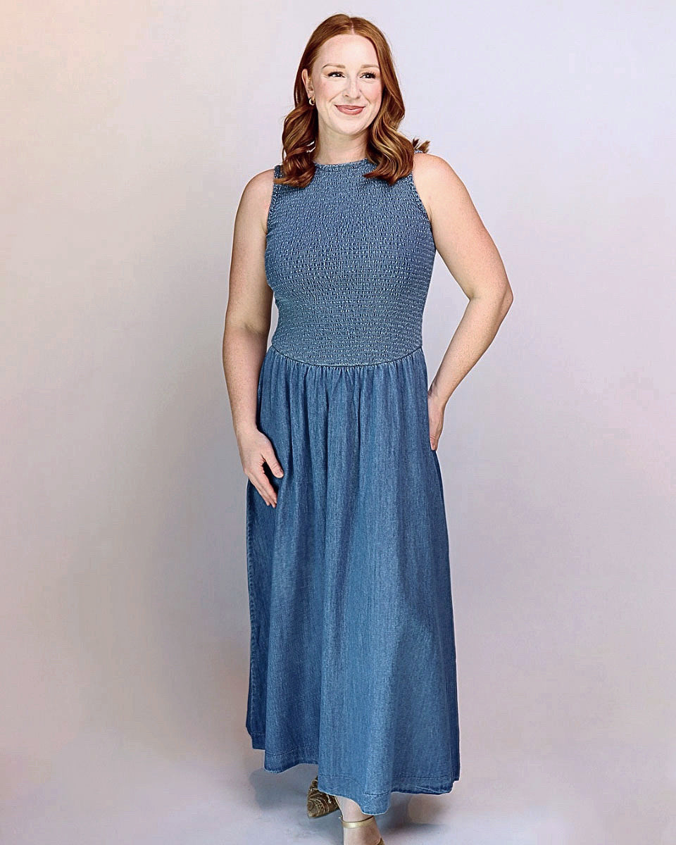 A woman modeling a sleeveless, high-neck, floor-length blue dress with a textured smocked bodice and a flowing skirt, set against a plain light gray studio background.