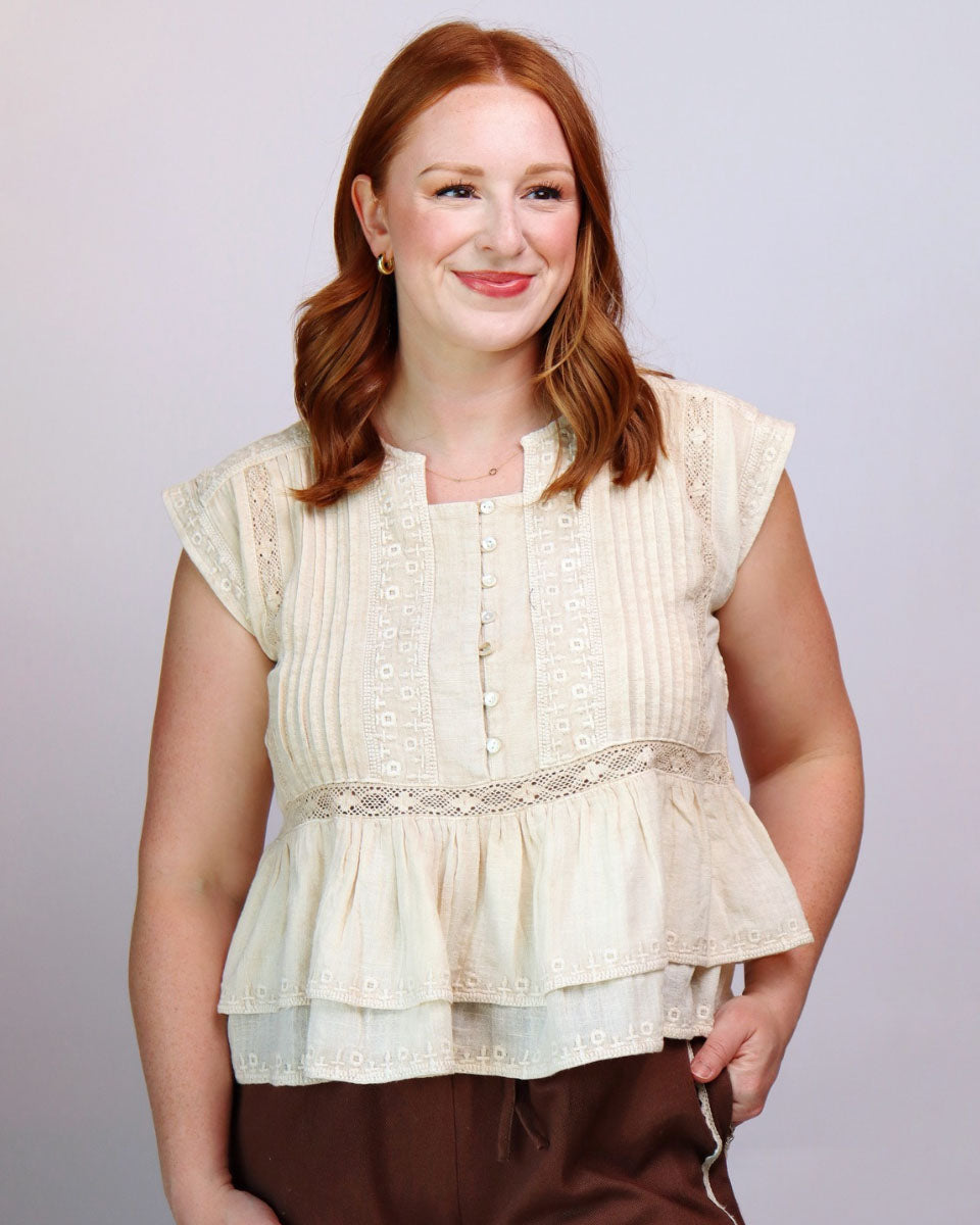 A woman with red hair wearing a cream-colored sleeveless peplum blouse with lace and button details, paired with brown pants, against a light gray studio background.