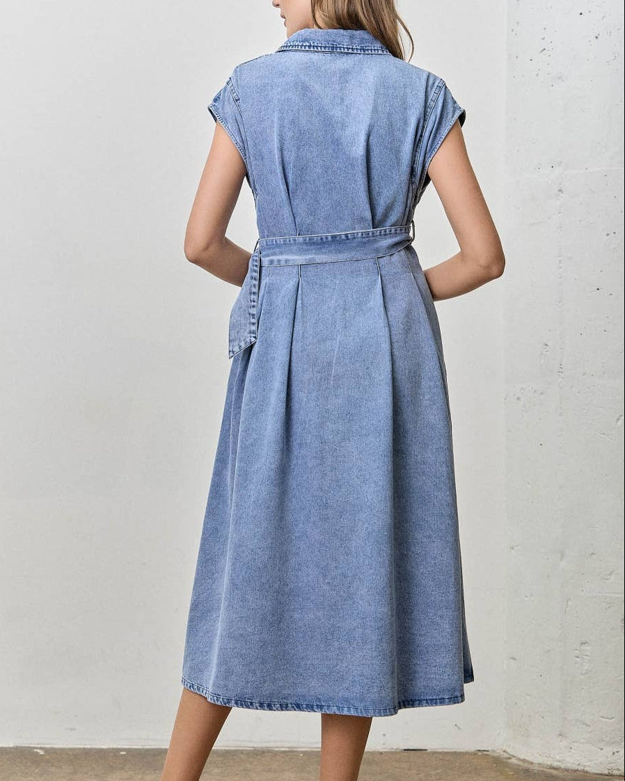 A woman wearing a light blue denim midi dress with cap sleeves and a tie waist, shown from the back in a studio setting.