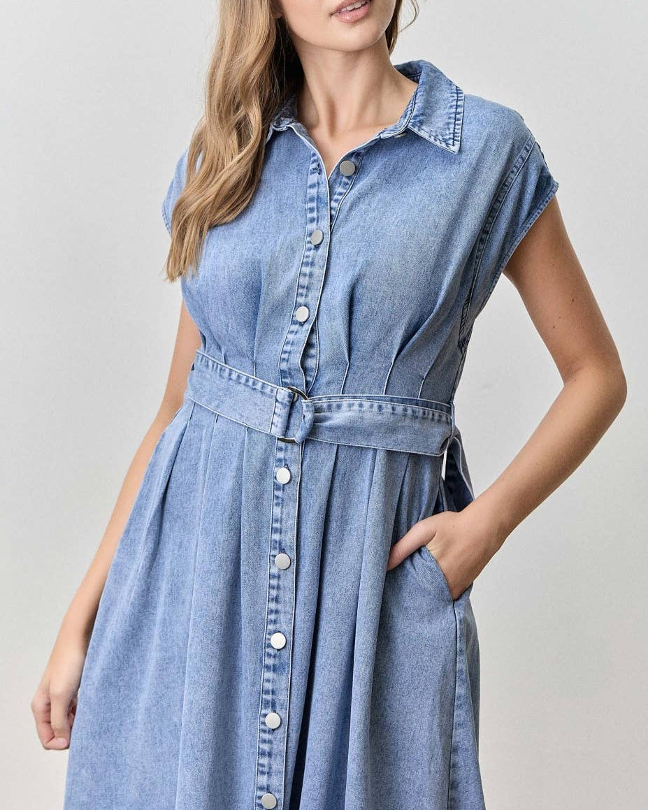 A woman modeling a light blue denim shirt dress with a button-down front, collar, and matching tie belt, photographed against a plain light gray studio background.