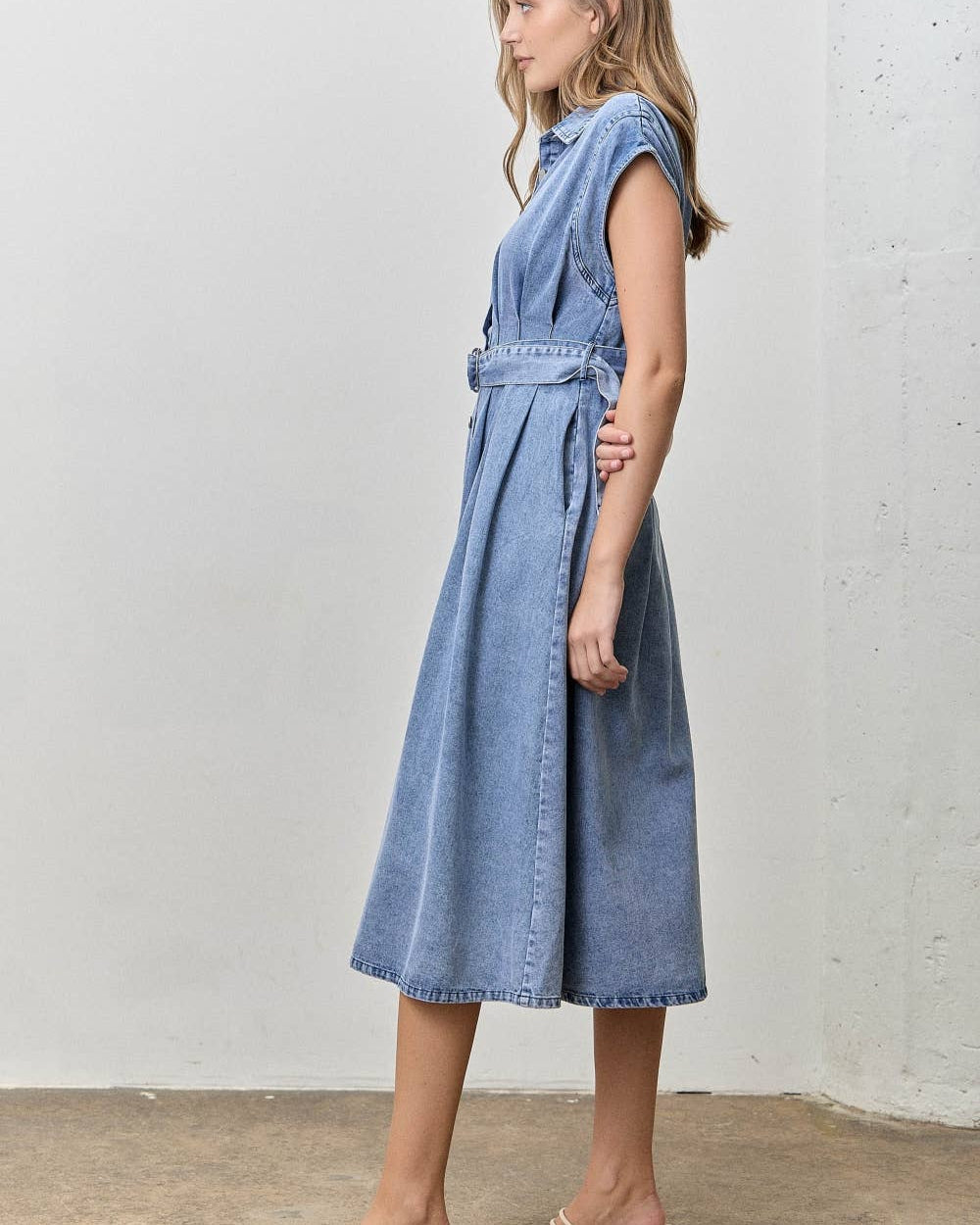 A woman wearing a light blue denim midi dress with a tie waist and sleeveless cap sleeves, paired with beige strappy block-heel sandals, standing against a textured off-white wall.