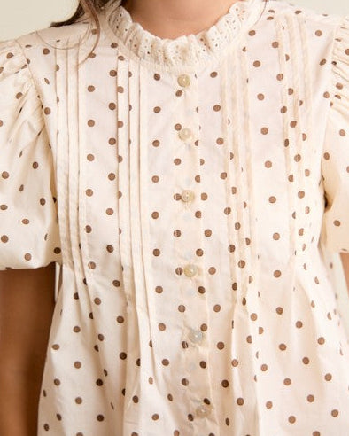 A woman wearing a cream-colored short-sleeve blouse with brown polka dots, featuring a ruffled high neckline and button-front detailing.
