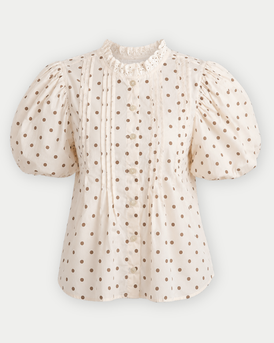 A cream-colored short-sleeve blouse with brown polka dots, featuring puffed sleeves, a ruffled high collar, and a button-up front.