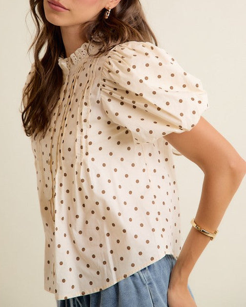 A woman wearing a cream-colored blouse with brown polka dots and puffed short sleeves, paired with light blue jeans, set against a plain beige background.
