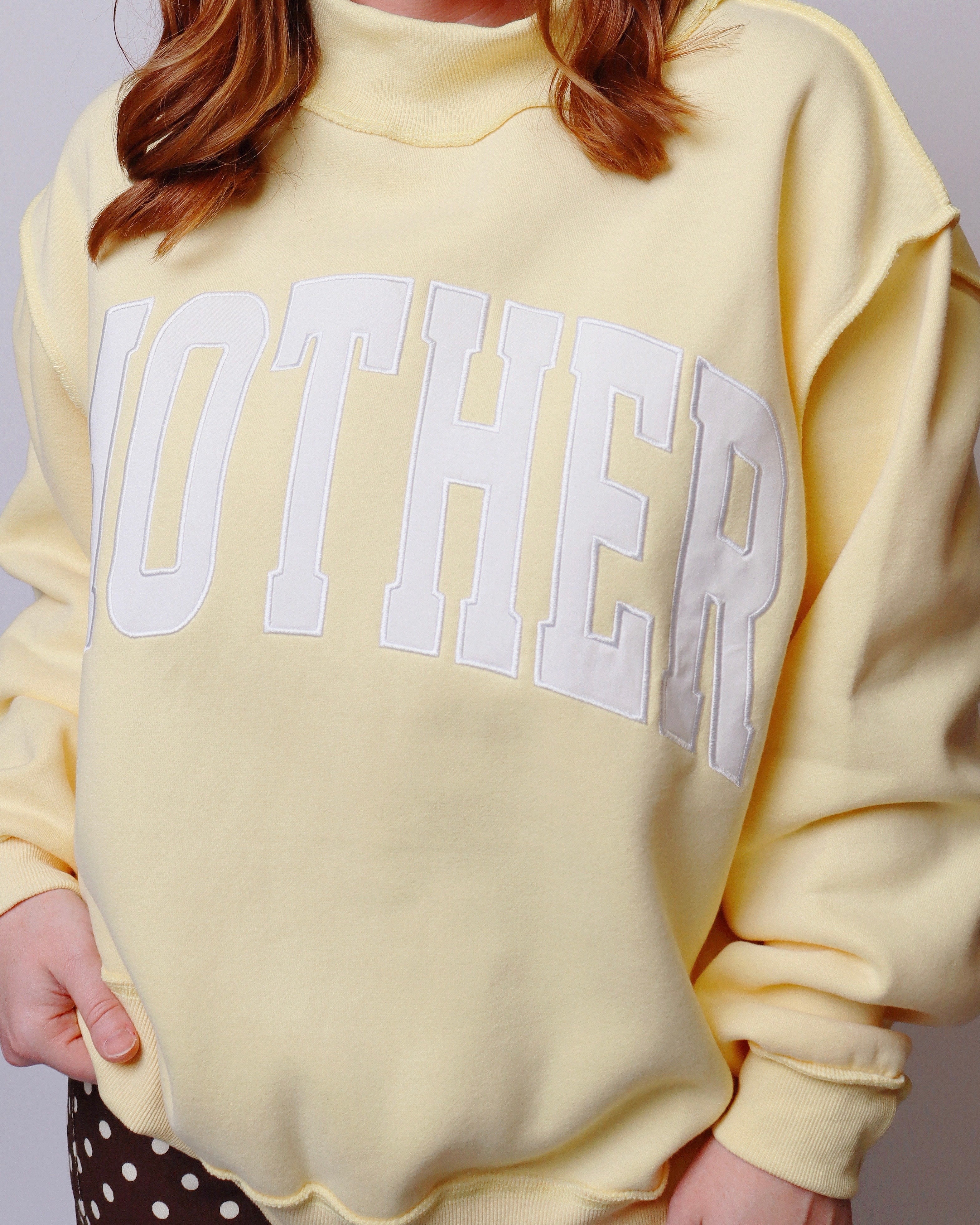A pale yellow sweatshirt with the word 'MOTHER' embroidered across the chest in large white varsity-style letters, shown on a model.
